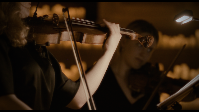Two female violinists playing in a dimly lit environment, focused on their sheet music, illuminated by stand lights, evoking a sense of concentration and teamwork.