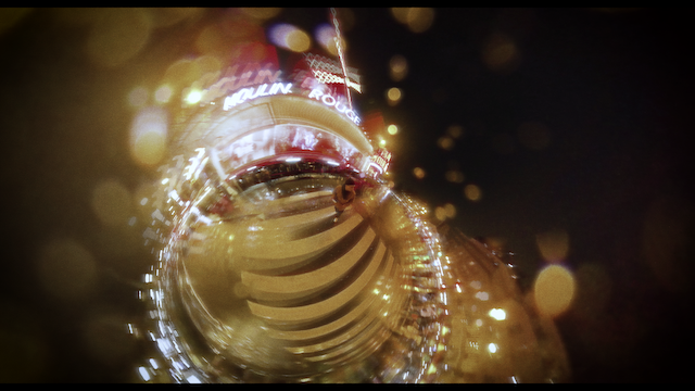 A creative, blurred shot capturing the bright lights and signage of the Moulin Rouge through a distorted glass that bends the light, creating a bokeh effect filled with warm tones.