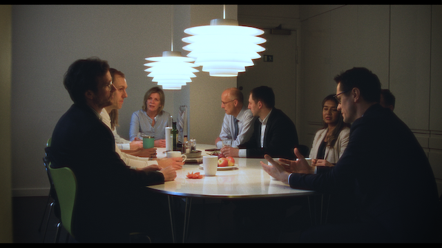 A business meeting in a modern office with Scandinavian-style chairs and pendant lighting. The table is surrounded by professionals engaged in a serious discussion.png
