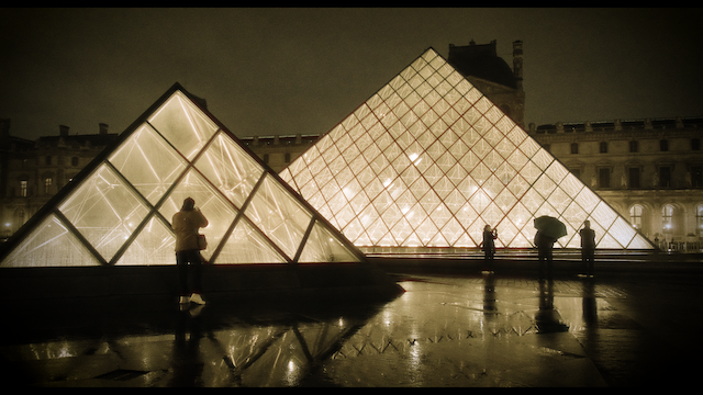 A nighttime view of the Louvre's glass pyramids, illuminated and reflecting on the wet ground, with silhouettes of people around them, creating a serene yet vibrant architectural spectacle.