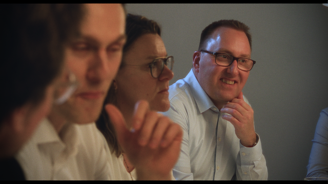 A close-up view of a business meeting with four people, focusing on a man in glasses smiling subtly at his colleagues during a discussion.png