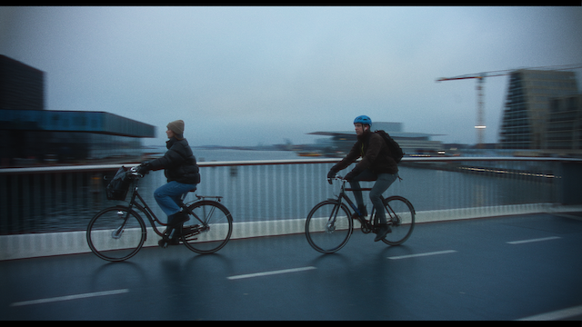 Two individuals cycling side by side on a bridge over a body of water in a city. The atmosphere is misty and the cyclists are wearing casual winter attire and helmets. Urban buildings and construction cranes are visible in the blurry background.png