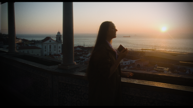 A woman stands on a balcony with her eyes closed, enjoying the sunset that casts a warm glow over the seaside cityscape behind her.png