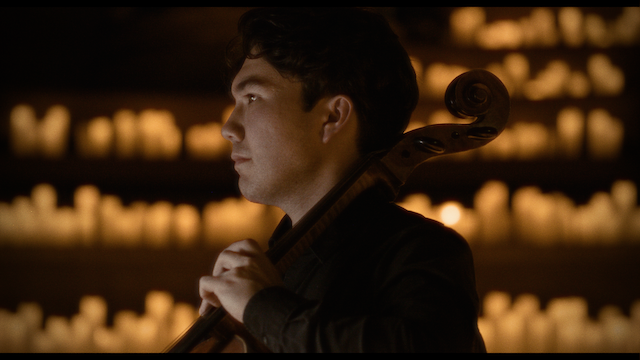 A male cellist, viewed from the side, holds his cello and bow with a focused expression, against a softly lit background suggesting an intimate performance setting.