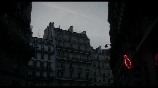 A moody, early evening shot of a Parisian street lined with traditional buildings, under a dusky sky with a lone neon sign glowing softly, evoking a sense of quiet urban life.