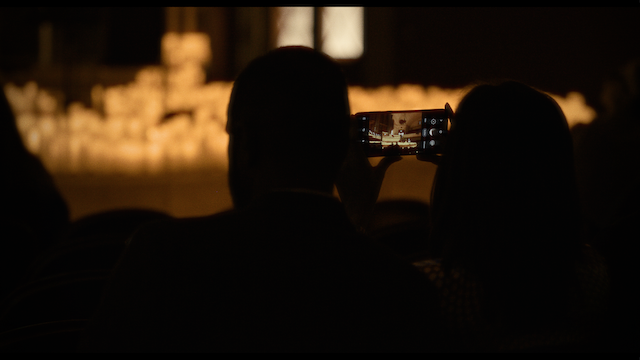 A silhouette of a couple sitting in an audience, capturing a performance on a smartphone, the stage bathed in a warm glow, enhancing the sense of a shared, intimate experience.
