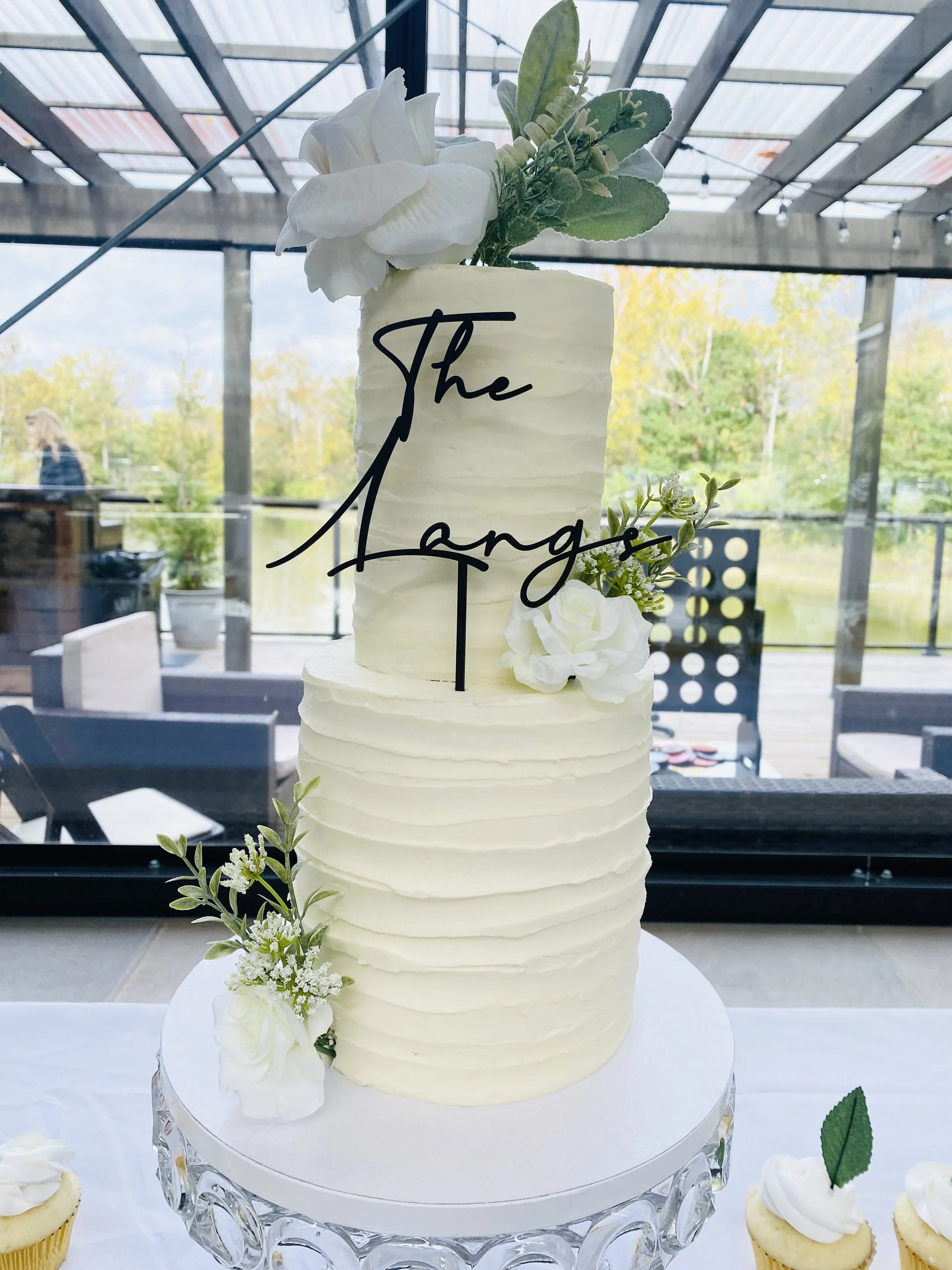 A tall wedding cake with white frosting and floral decorations, topped with a black monogram cake topper that reads 'The Mangos', placed on a glass cake stand. There are cupcakes with white frosting and a green leaf on a table in front of the cake, w