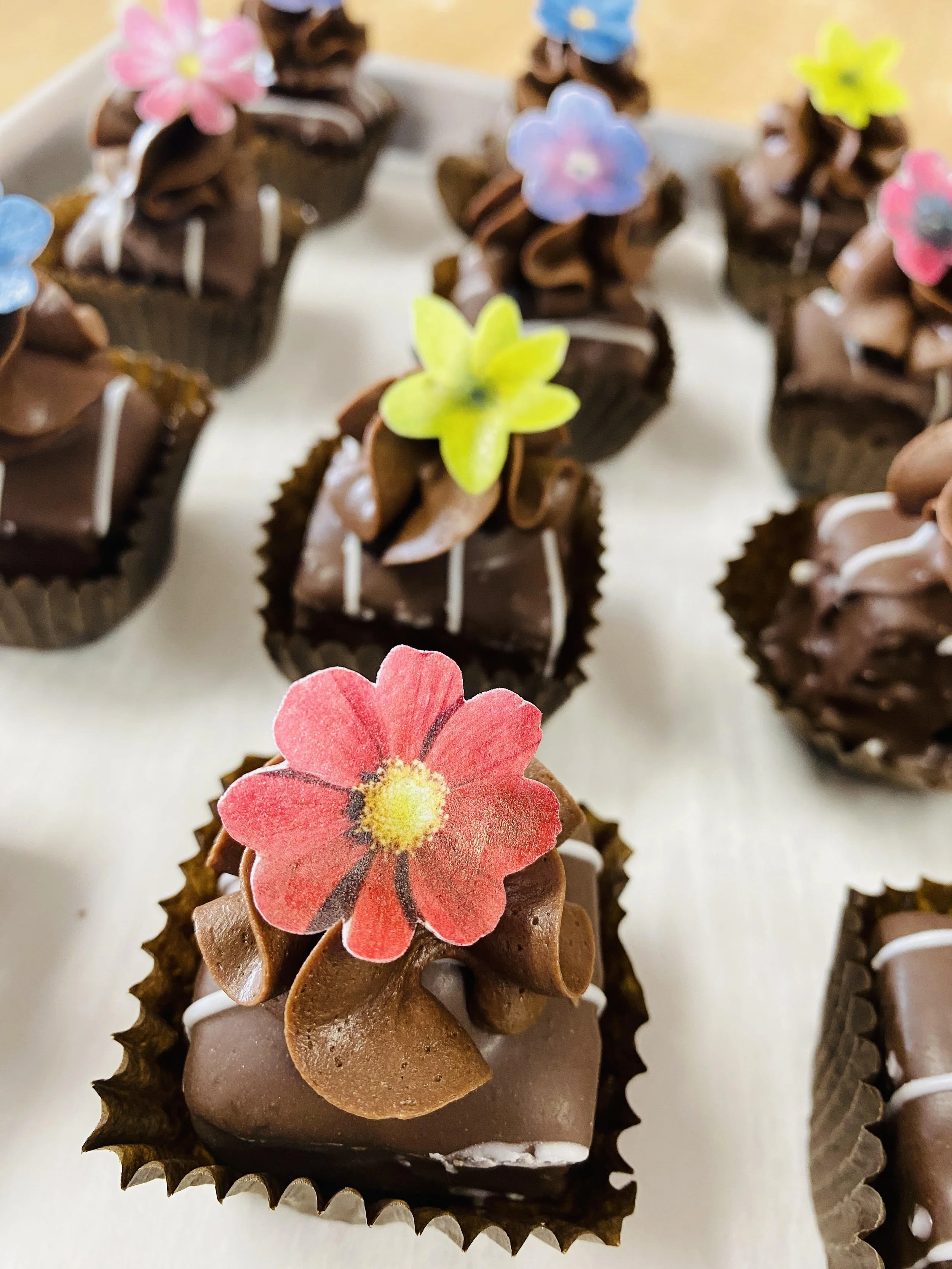 Assorted chocolate cupcakes decorated with colorful edible paper flowers on a white surface.