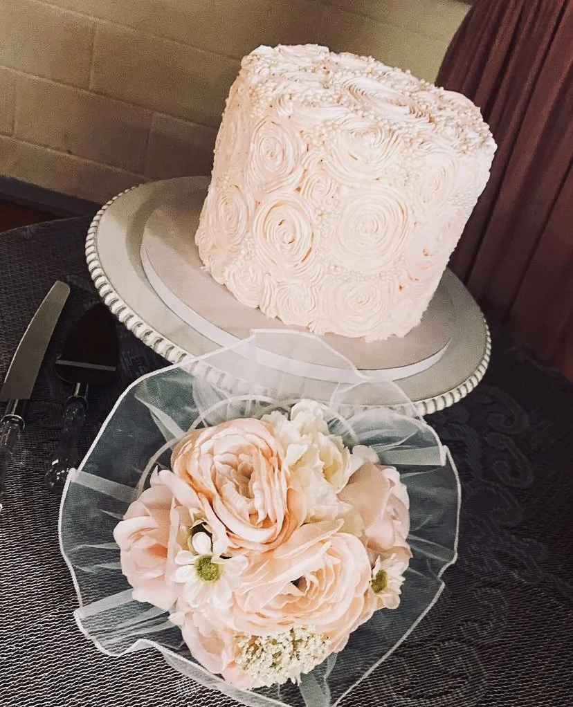 A pink, square wedding cake with rosette piping decoration sits on a silver cake board. In front of it, a bouquet of large, soft pink and white flowers, wrapped in sheer material, rests on a black tablecloth.