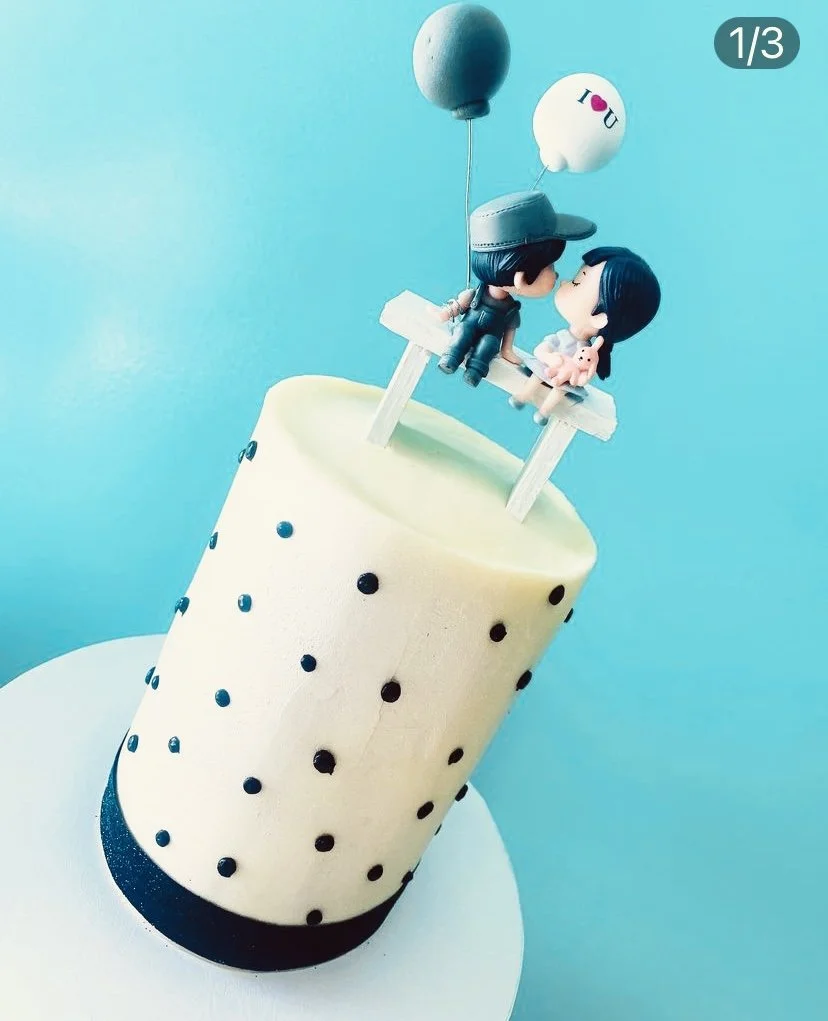 A cake designed to look like a bubble tea drink with two cartoon figures sitting on top, one with a hat and the other holding a small pig, with balloons above them. The background is light blue.
