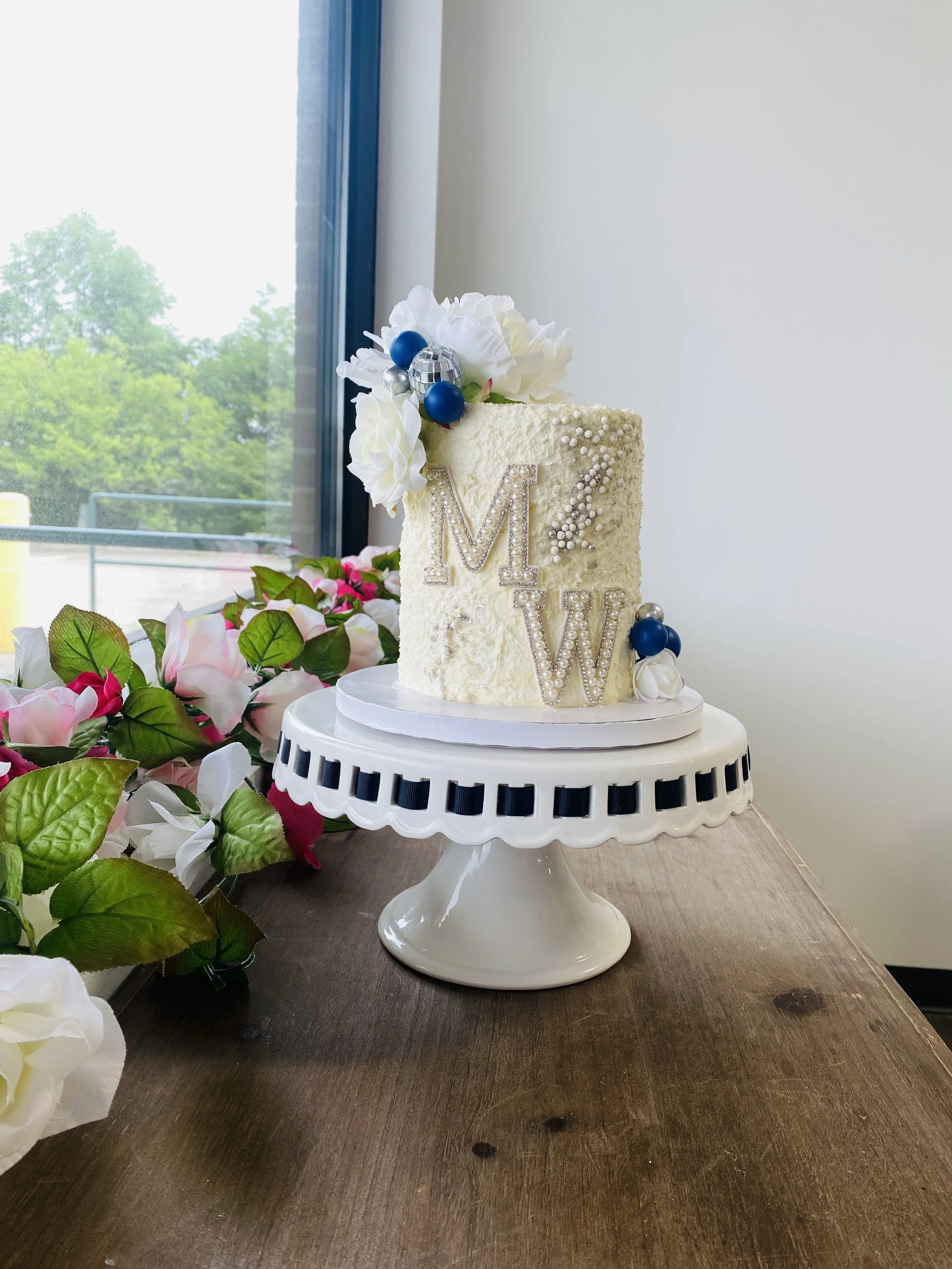 Decorative wedding cake with white icing, topped with white flowers and blue and silver balls, placed on a white cake stand with black and white trim, on a wooden table with pink and white flowers nearby, against a window showing green trees.