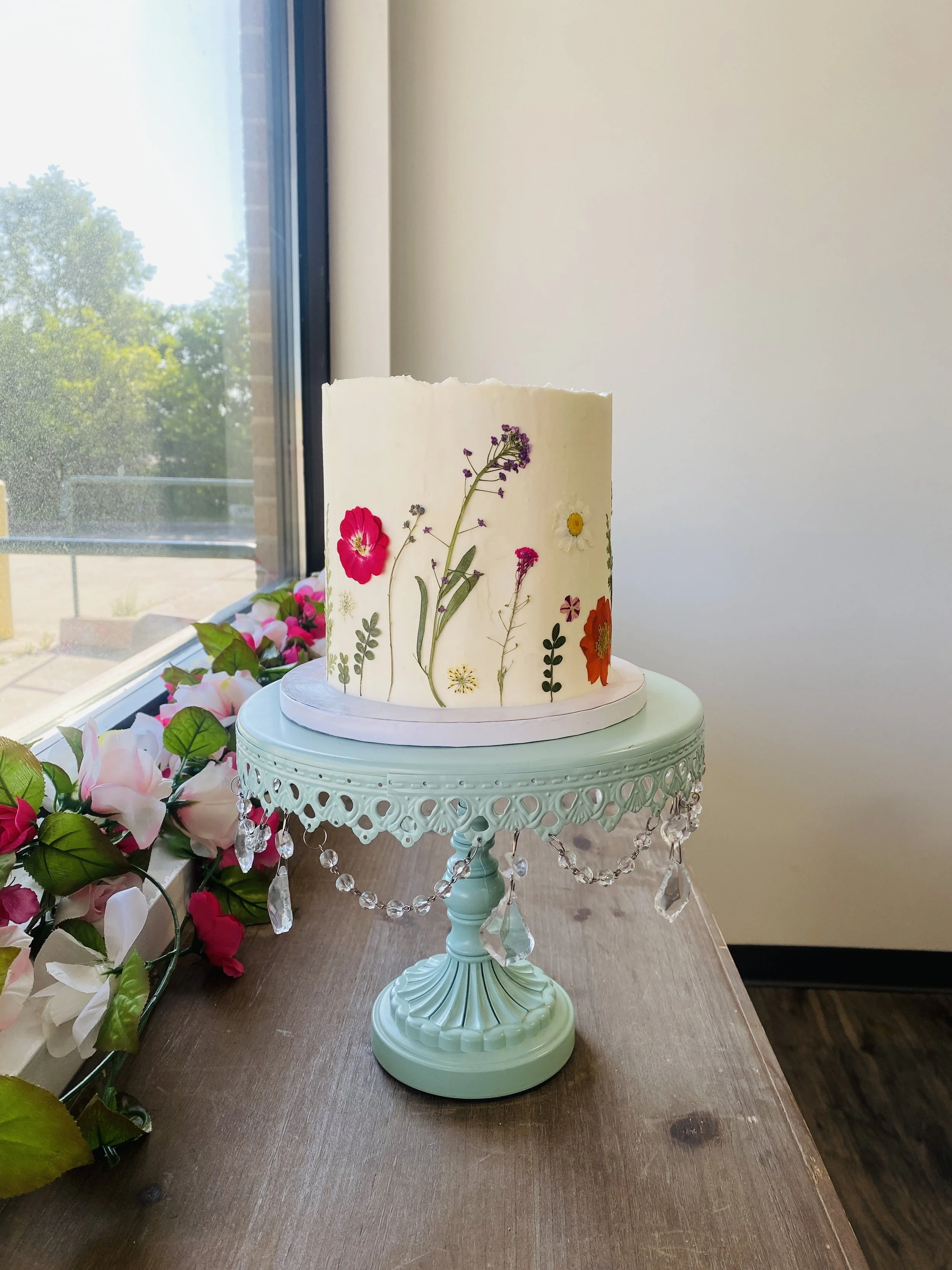 A white cylindrical cake decorated with pressed flowers, placed on a mint green ornate cake stand with hanging beads, next to pink and white artificial flowers near a window.