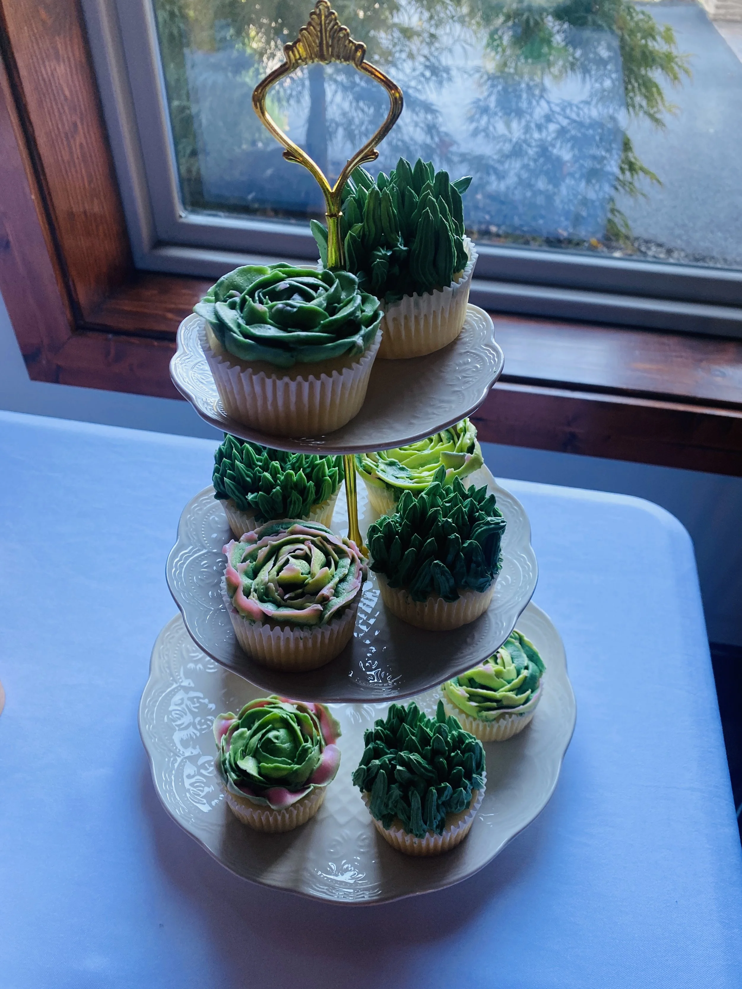 Cupcakes decorated to look like succulent plants on a three-tiered white serving platter near a window.