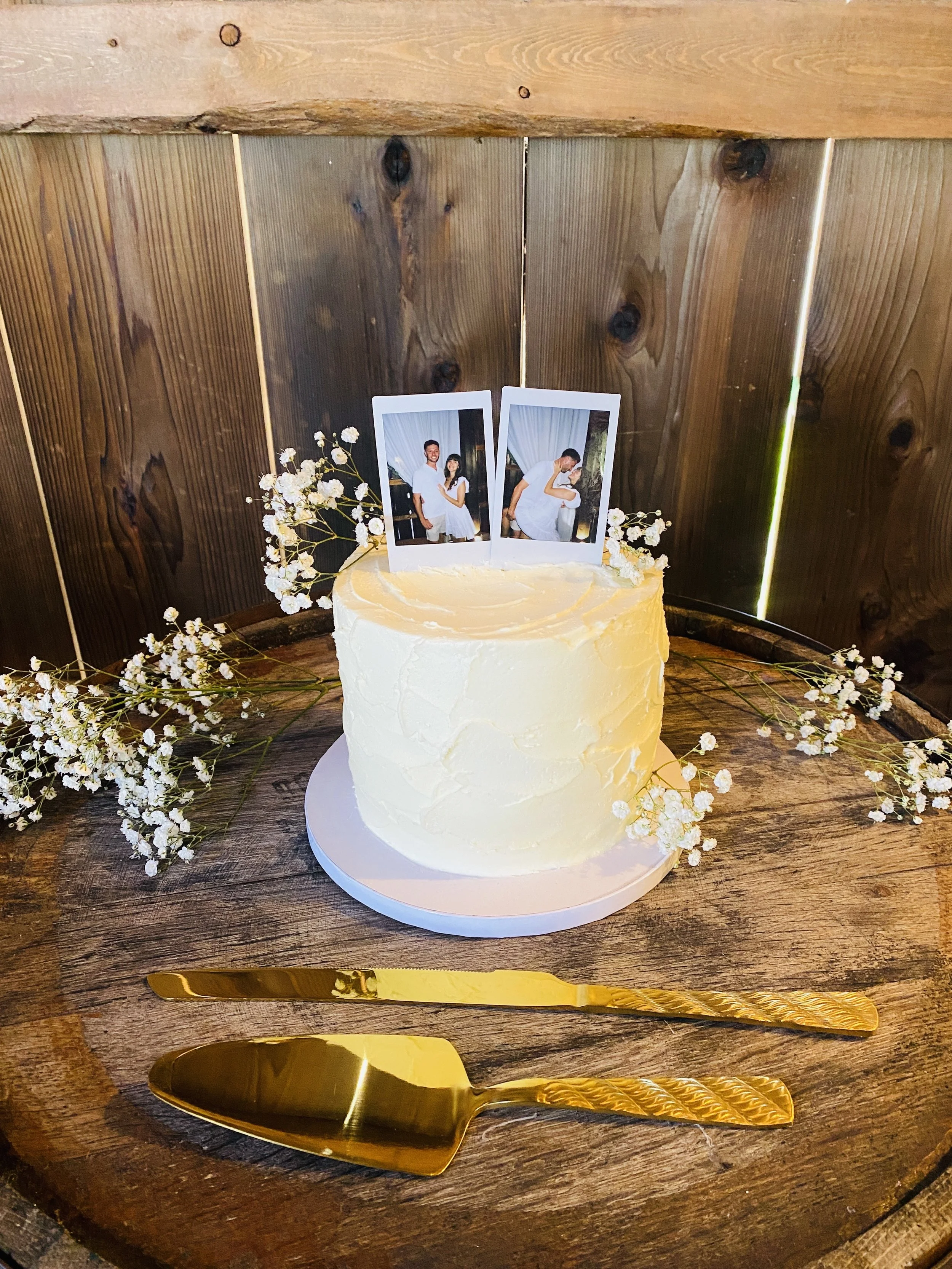 A wedding cake decorated with white flowers and two instant photos of a couple, placed on a rustic wooden surface with gold cake server and knife.