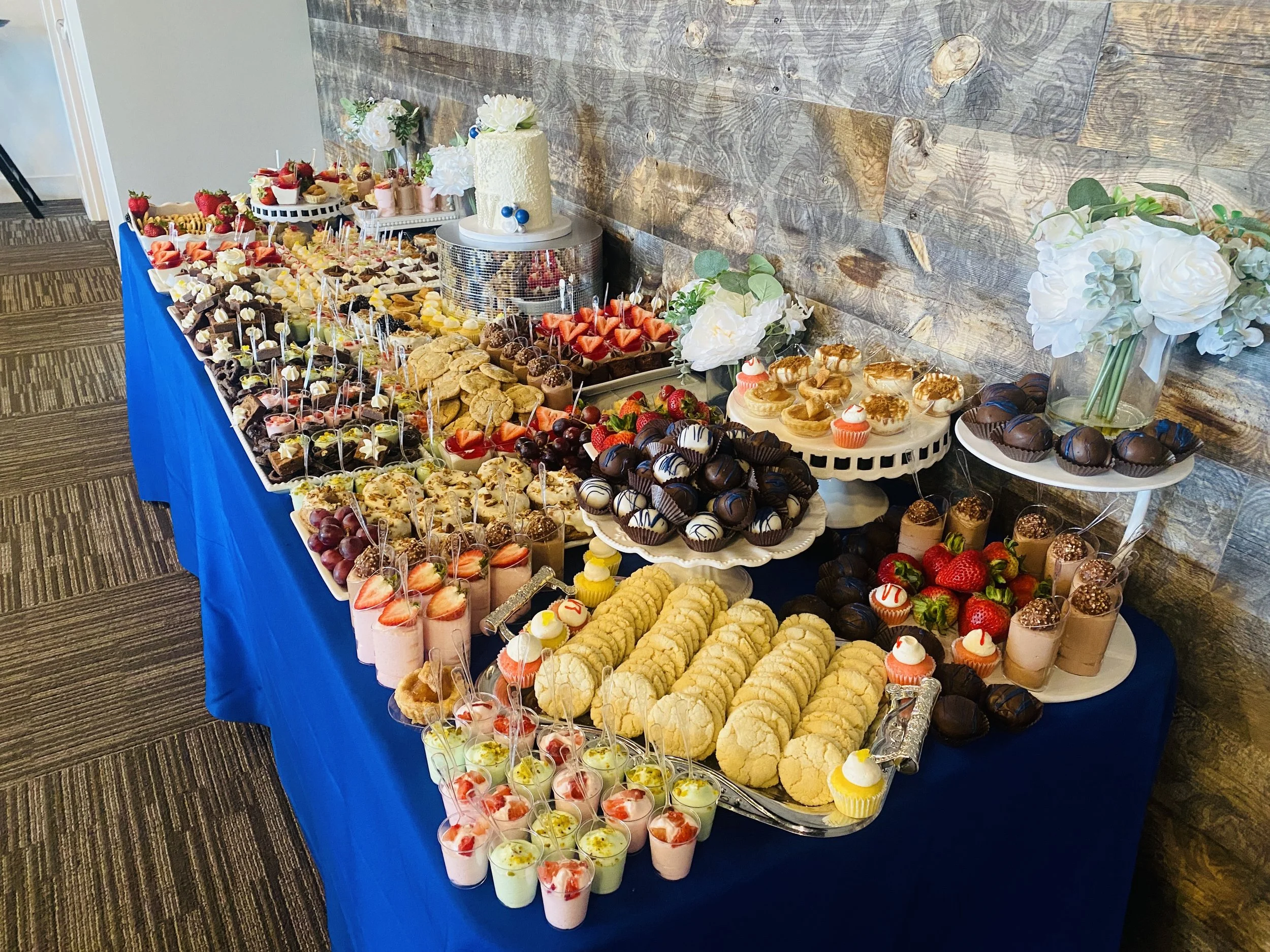 Dessert table with cupcakes, cookies, strawberries, chocolate truffles, and other sweets on a blue tablecloth, against a wooden wall.