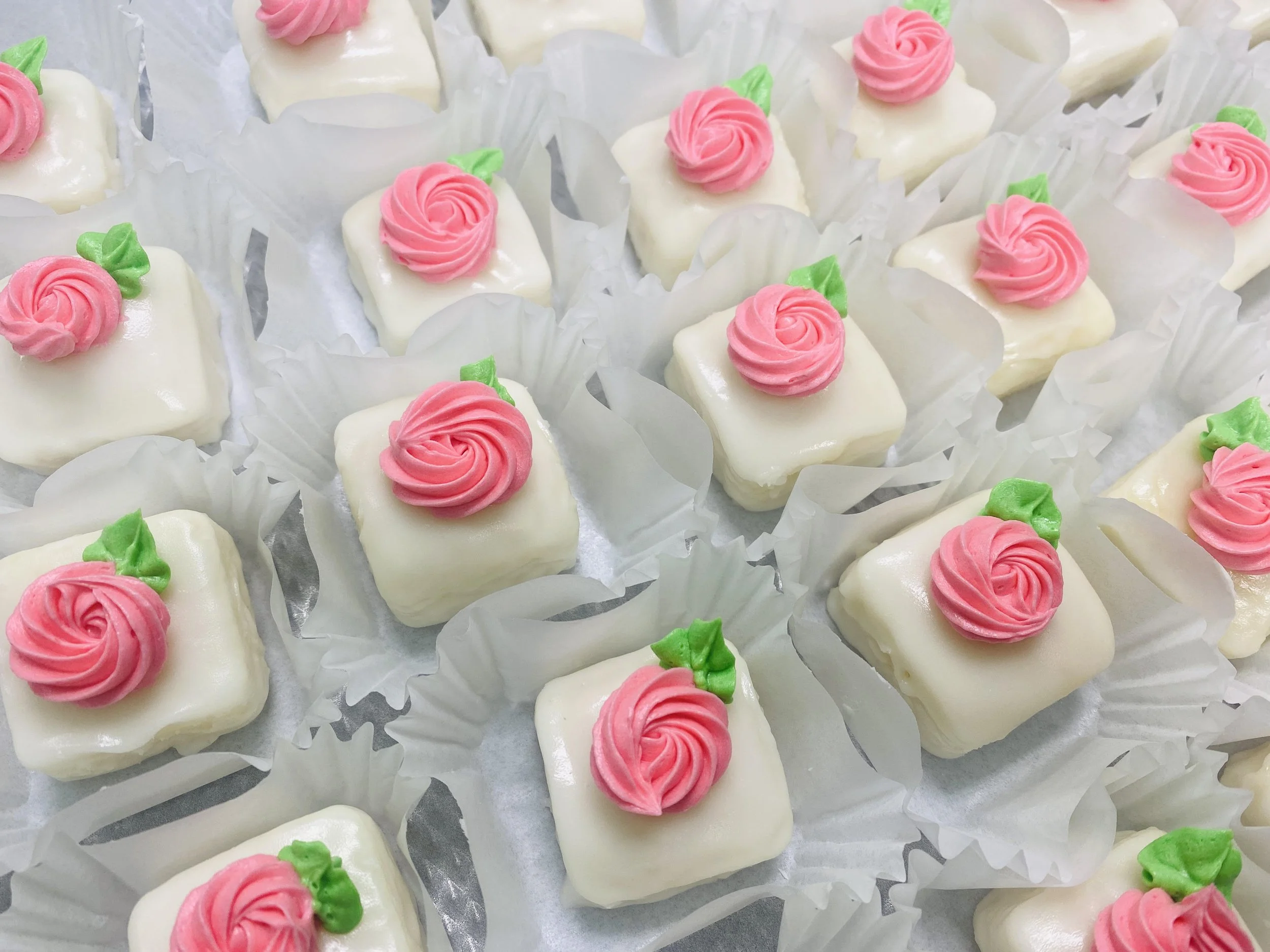 Row of small square cakes covered in white icing, decorated with pink icing roses and green leaves, each in paper cupcake liners.