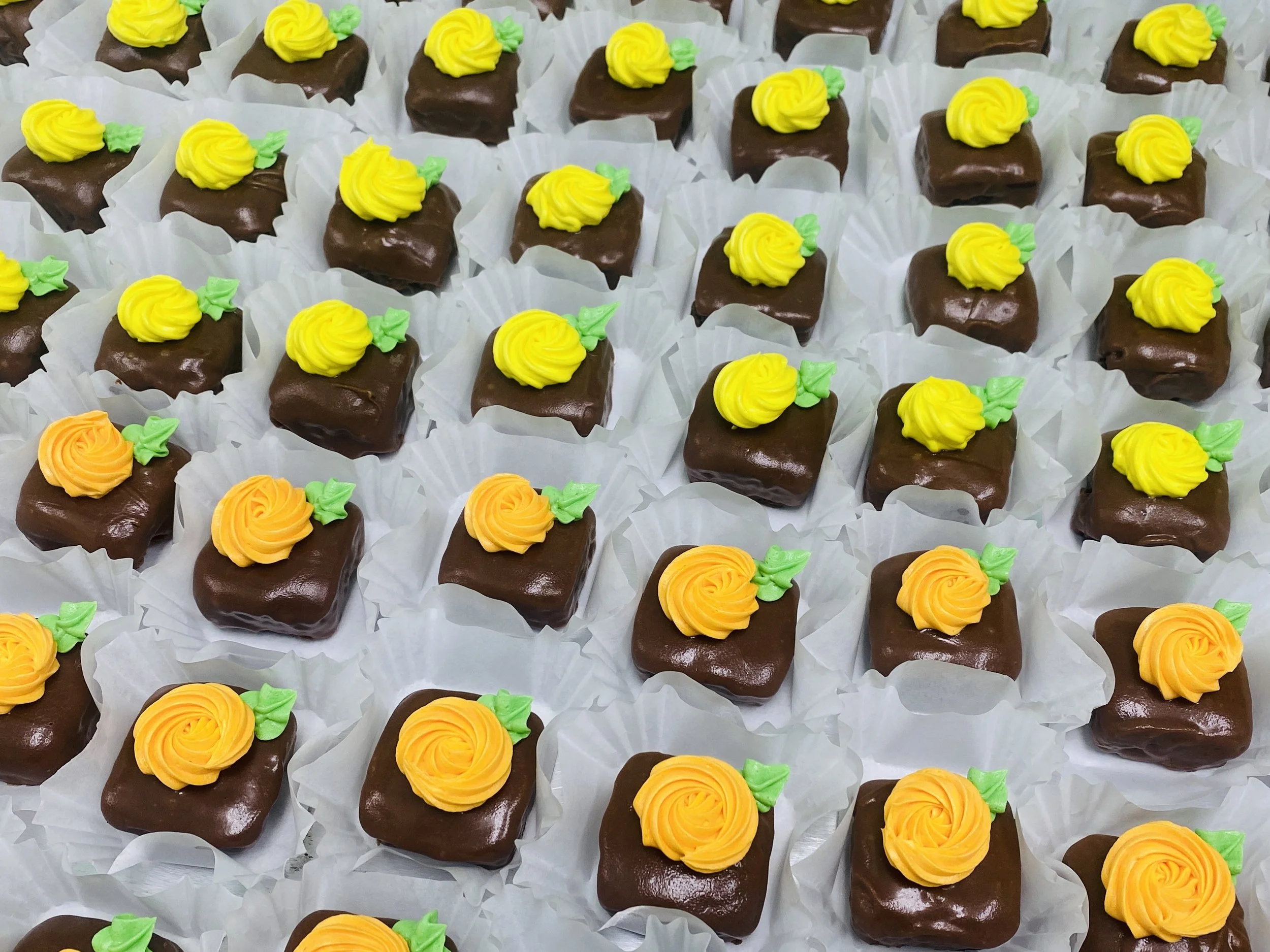 A large tray of square chocolate petit fours decorated with yellow and orange buttercream rosettes and small green buttercream leaves, each placed on individual white paper cups.