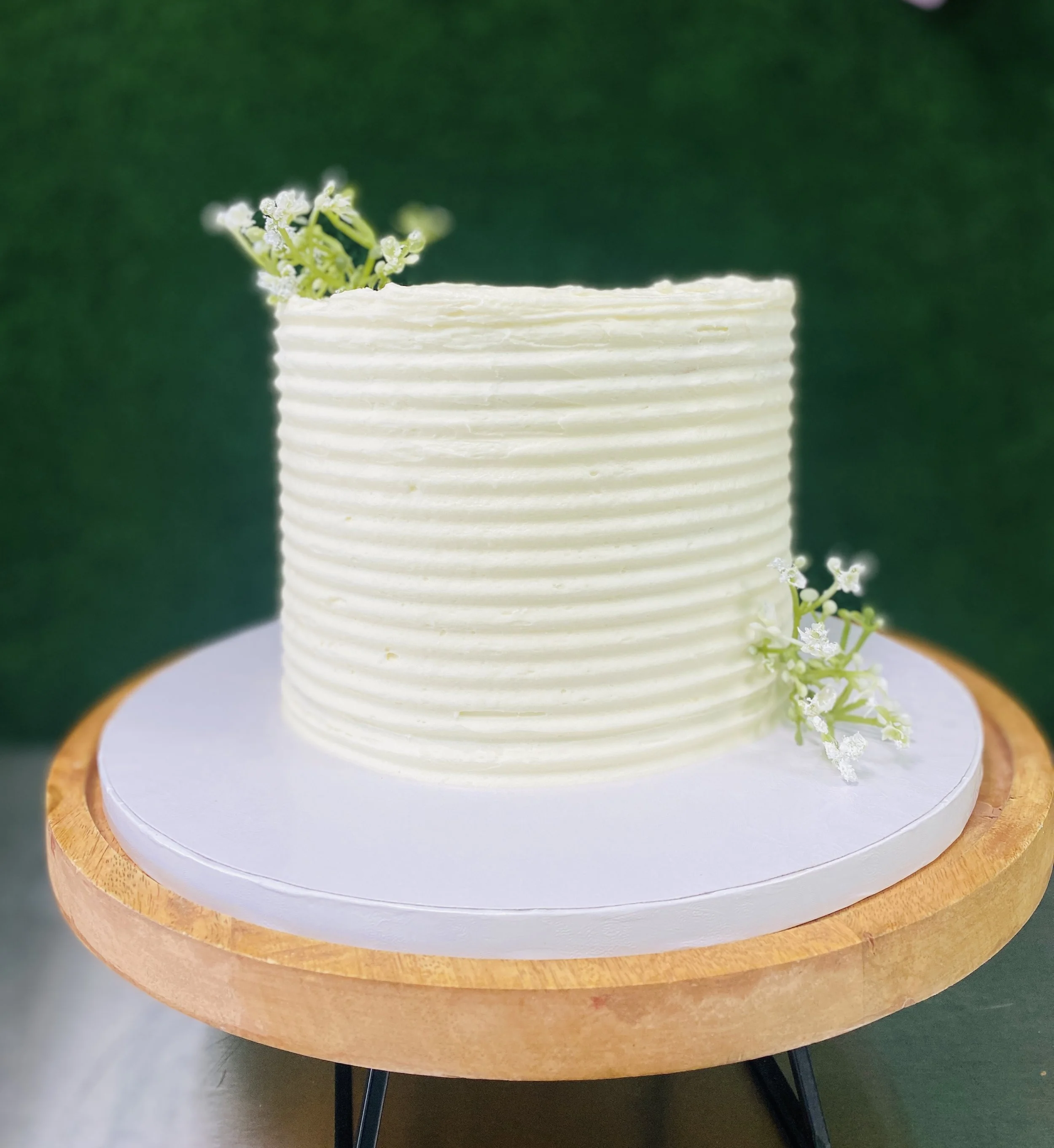 White frosted cake with textured horizontal lines, decorated with small white flowers, placed on a white cake board on a wooden cake stand with black legs, against a green background.