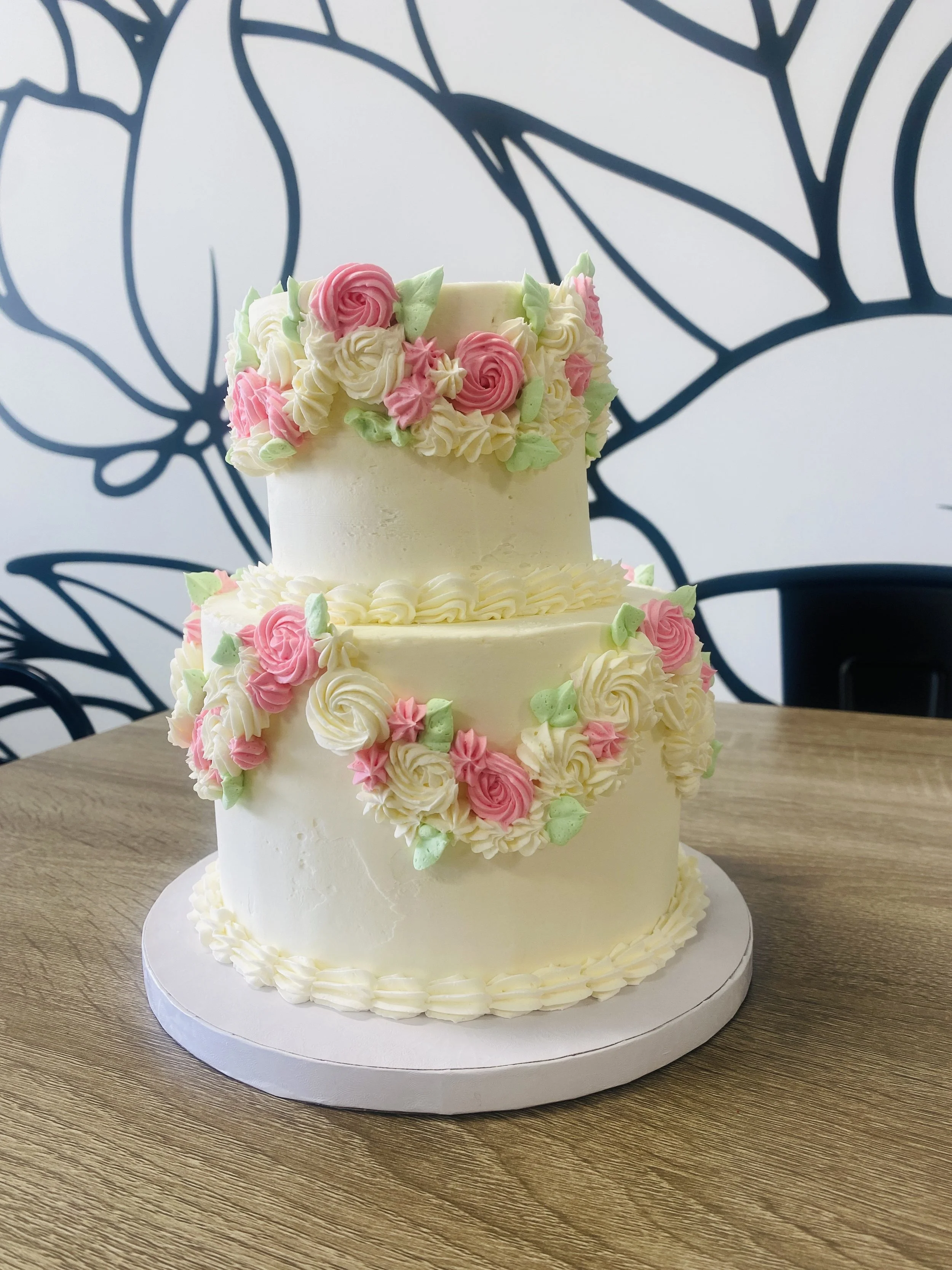 A three-tiered white wedding cake decorated with pink and white buttercream roses, green leaves, and piped borders, placed on a wooden table.