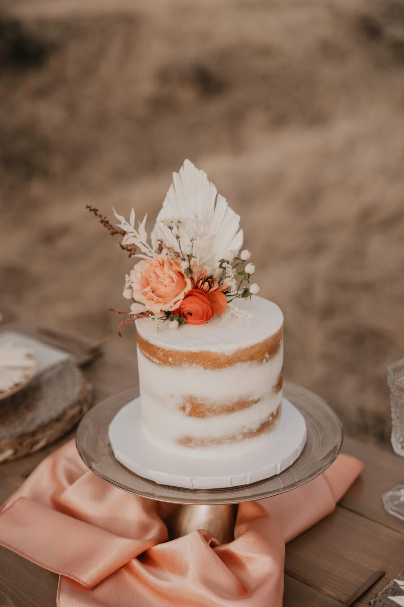 A small, semi-naked cake decorated with white icing and fresh peach-colored flowers, topped with a white leaf and small white berries, on a silver cake stand.