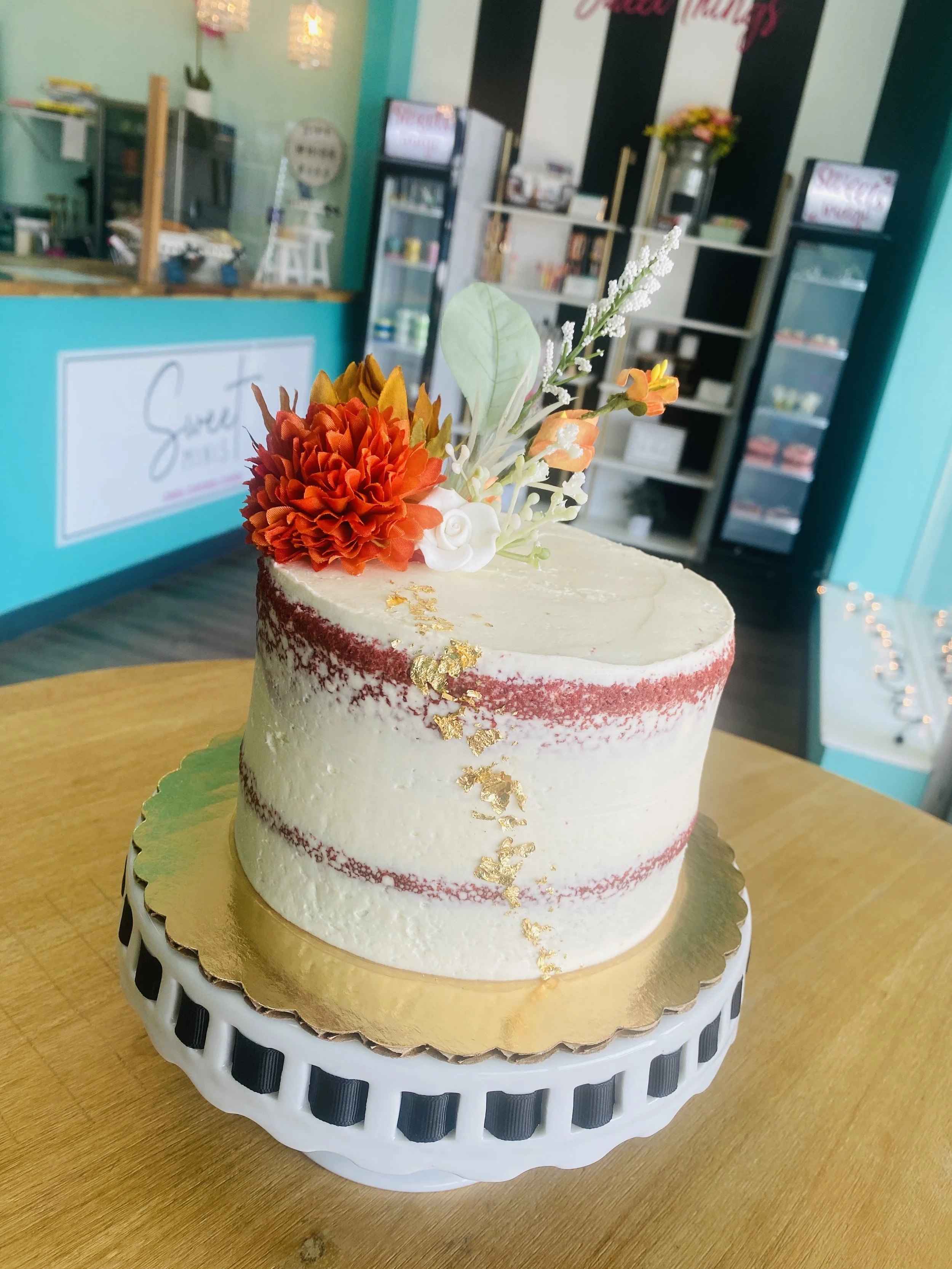A decorated cake with white and red frosting, gold leaf accents, and a floral topper with orange and white flowers, on a gold cake board and white stand, inside a bakery shop.