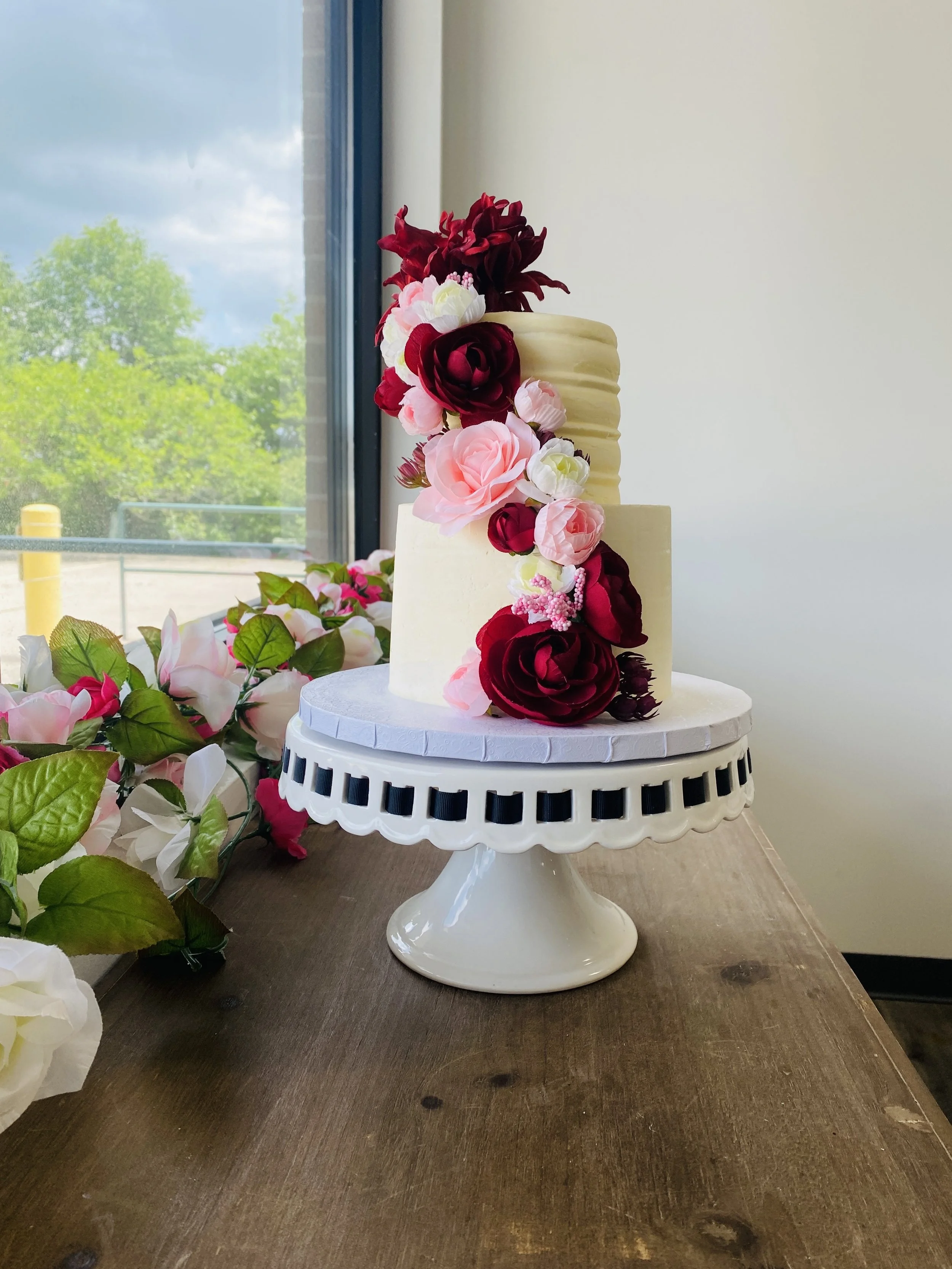 A two-tier white wedding cake decorated with pink and burgundy flowers, placed on a white cake stand on a wooden table near a window with greenery outside.