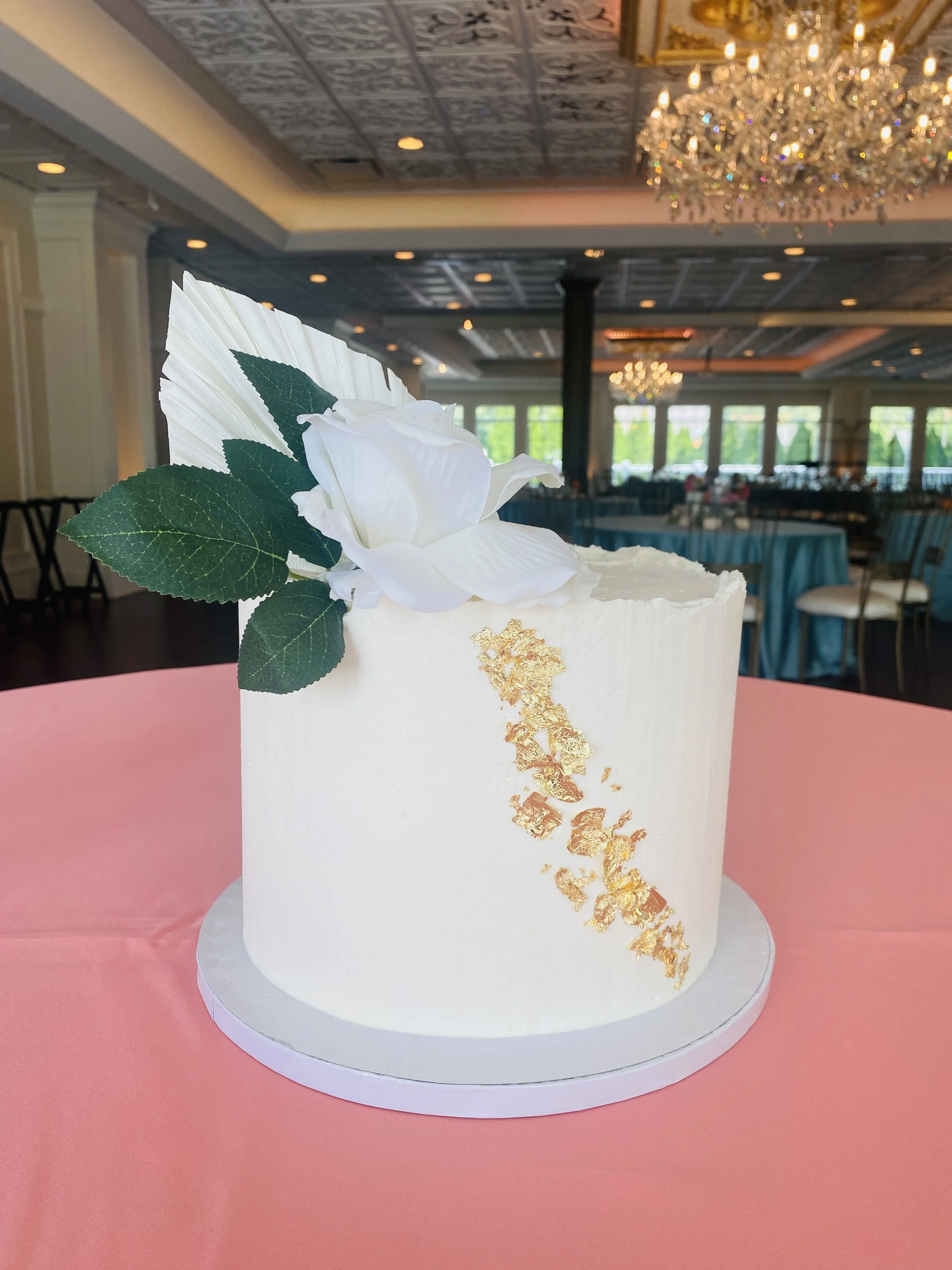 White wedding cake with a white flower and green leaves. Gold foil accents on the side. The cake is on a pink tablecloth in a decorated banquet hall.
