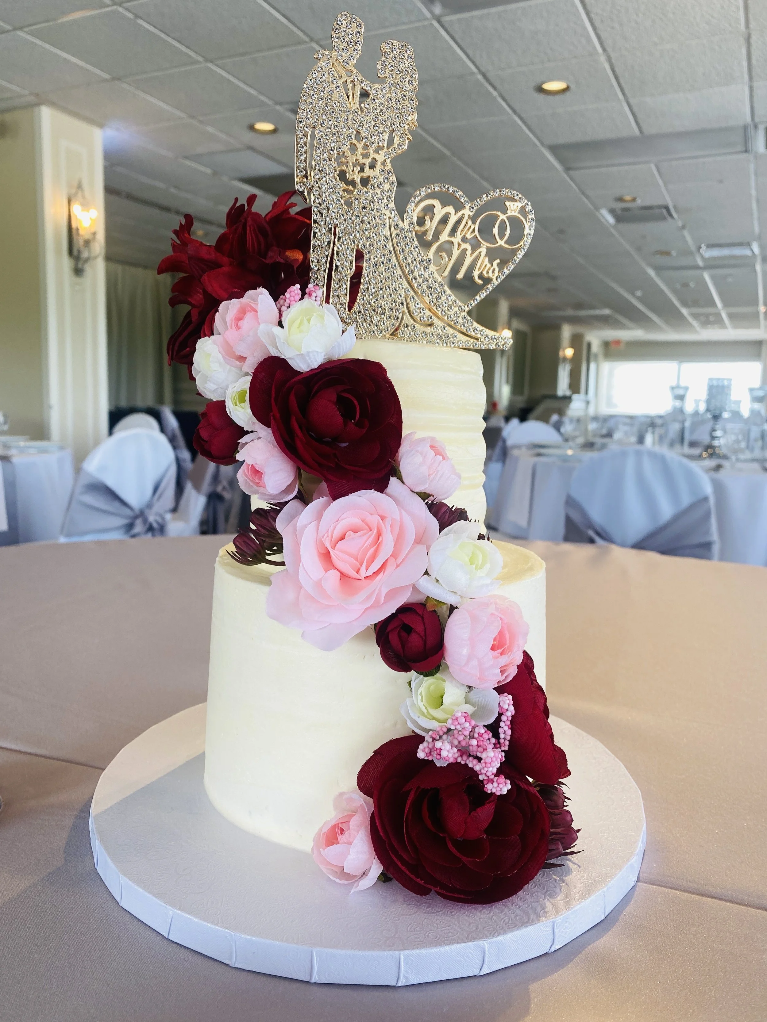 A two-tier wedding cake decorated with pink and deep red roses, topped with a glittery silhouette of a newlywed couple and a heart-shaped topper that says 'Mr & Mrs'. the cake is on a white round base, placed on a beige tablecloth in an event hall.