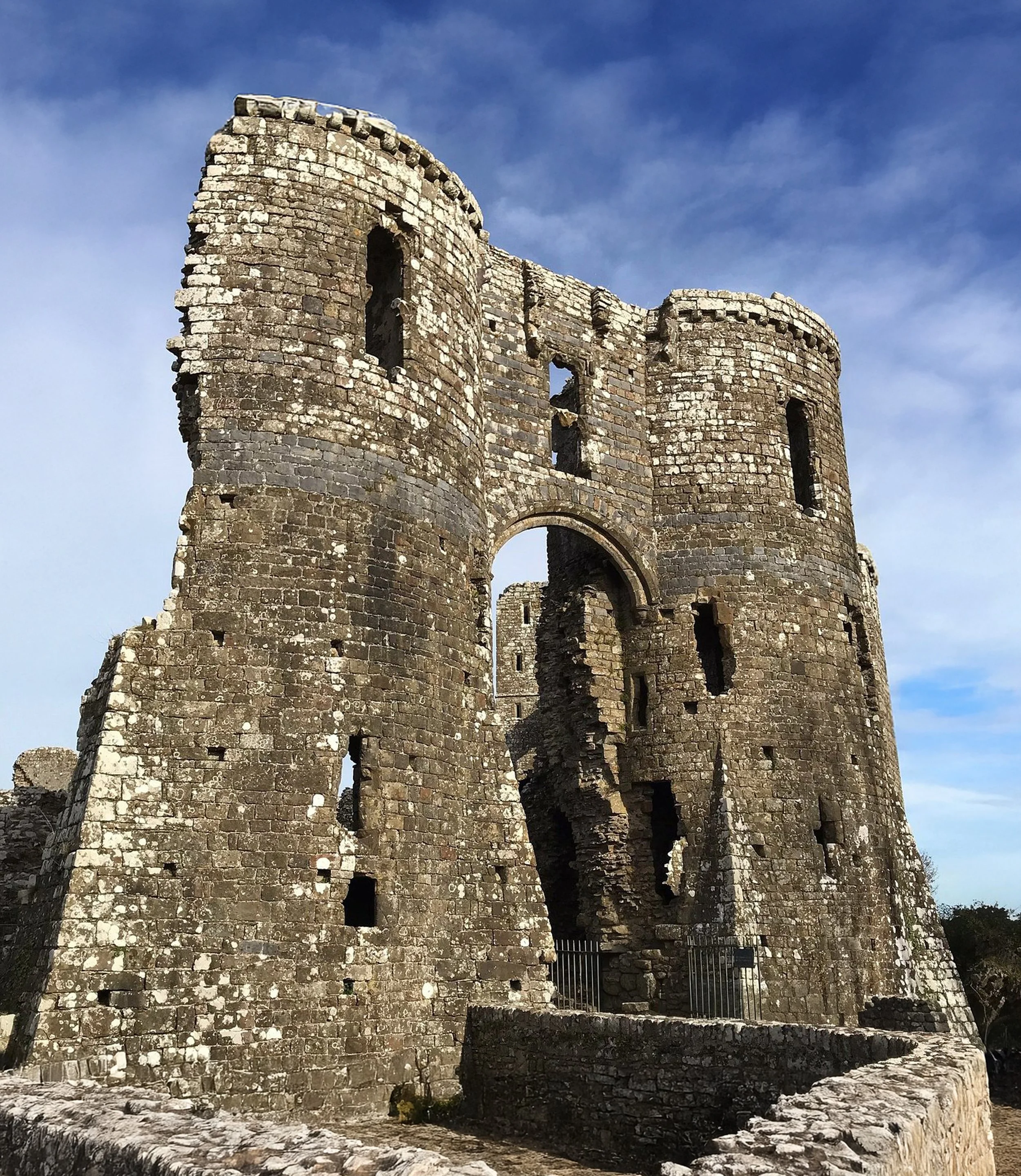 A view of an ancient stone castle tower with a rounded top, surrounded by greenery and trees, partially obscured by a stone wall in the foreground.