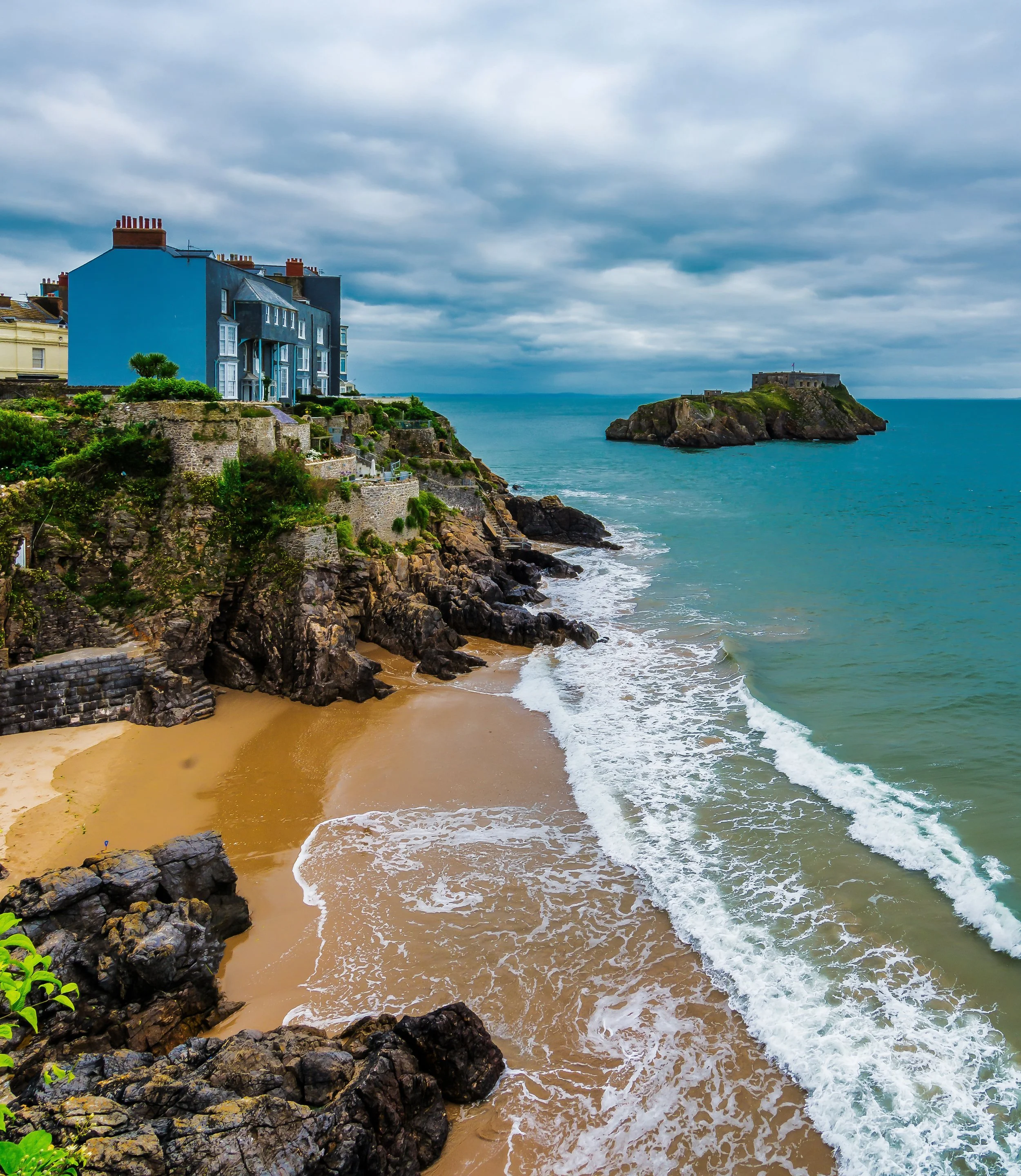 A harbor with multiple boats docked along piers, a sandy beach behind a stone wall, and colorful buildings on a hill overlooking the harbor, under a blue sky.