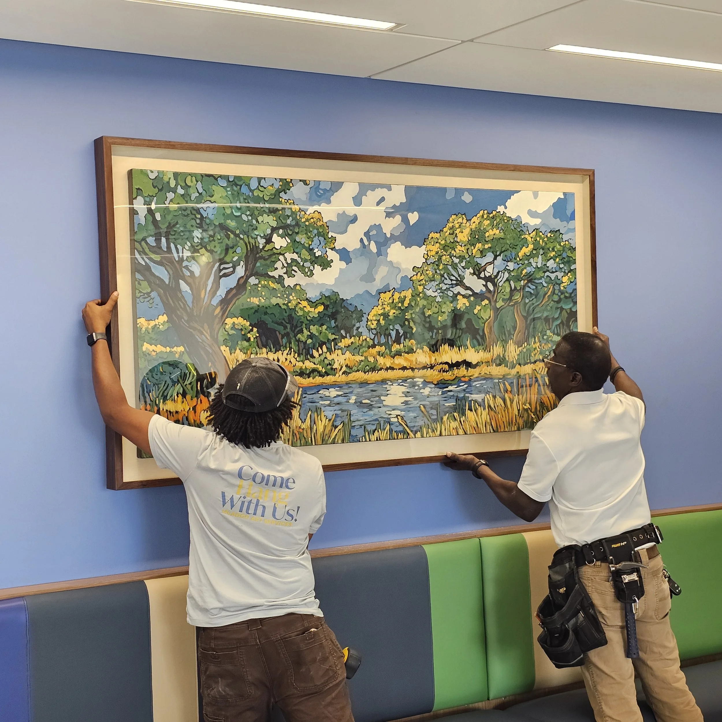 Two workers hanging a large colorful landscape painting on a blue wall inside a building.