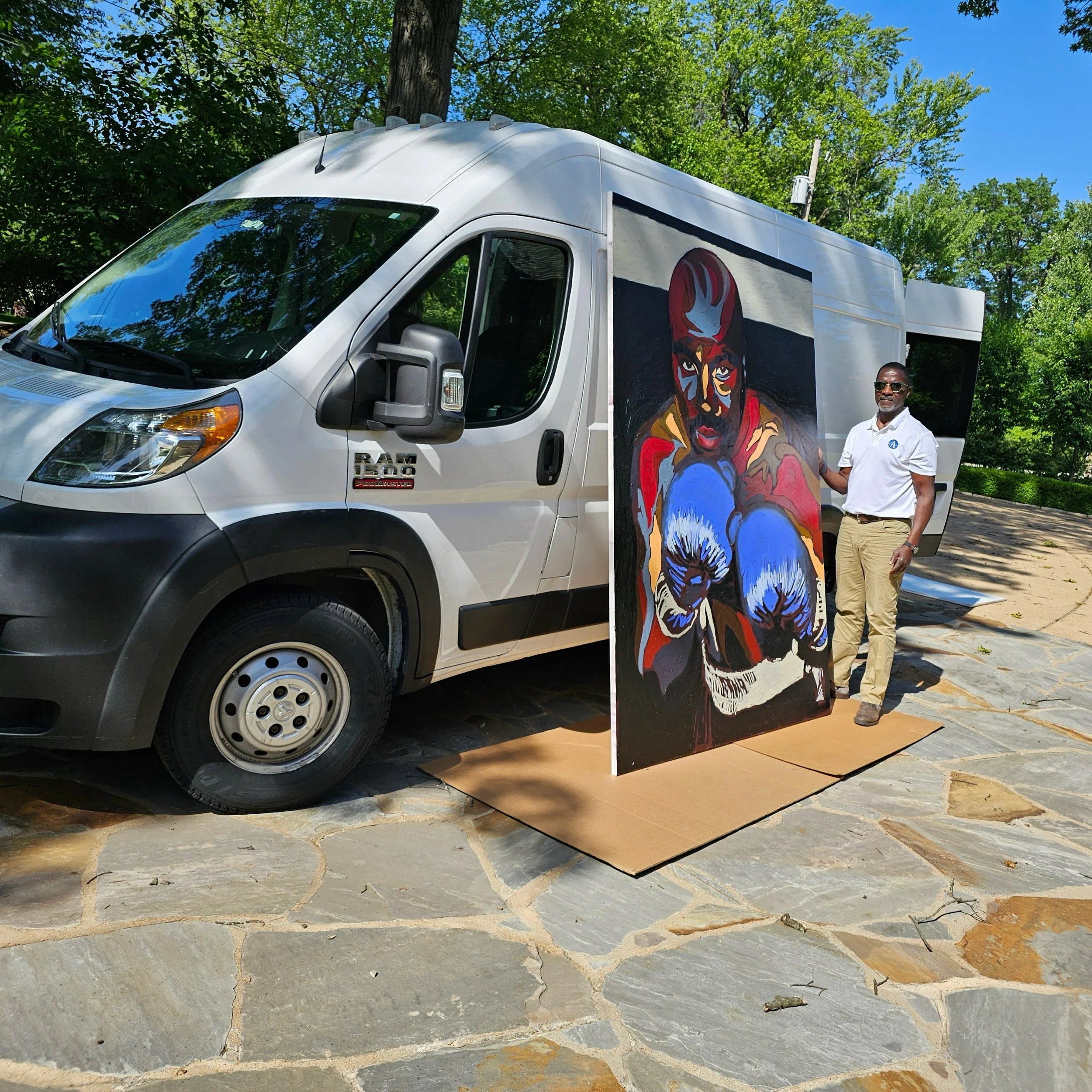 A man standing beside a decorated van with mural artwork on its side, featuring a boxer with gloves and a hooded figure, outdoors with trees and a clear blue sky.