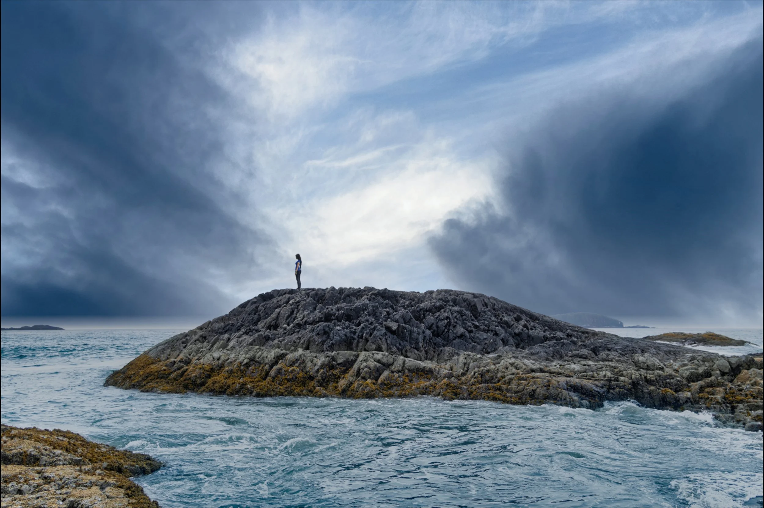A person standing alone on a rocky island surrounded by ocean, under a cloudy sky.
