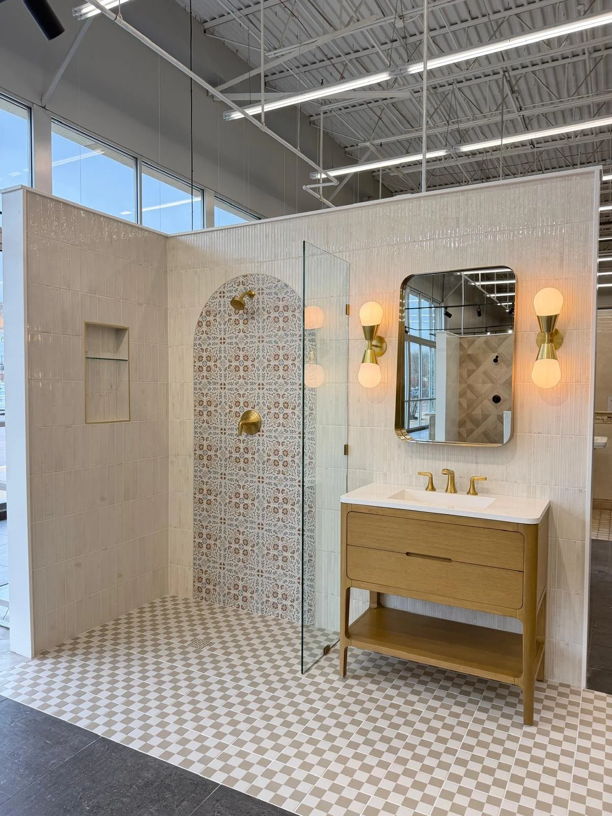 A bathroom display featuring a walk-in shower with patterned tiles, a mirror, a modern vanity with gold fixtures, and wall sconces with white shades, all within a home improvement store setting.