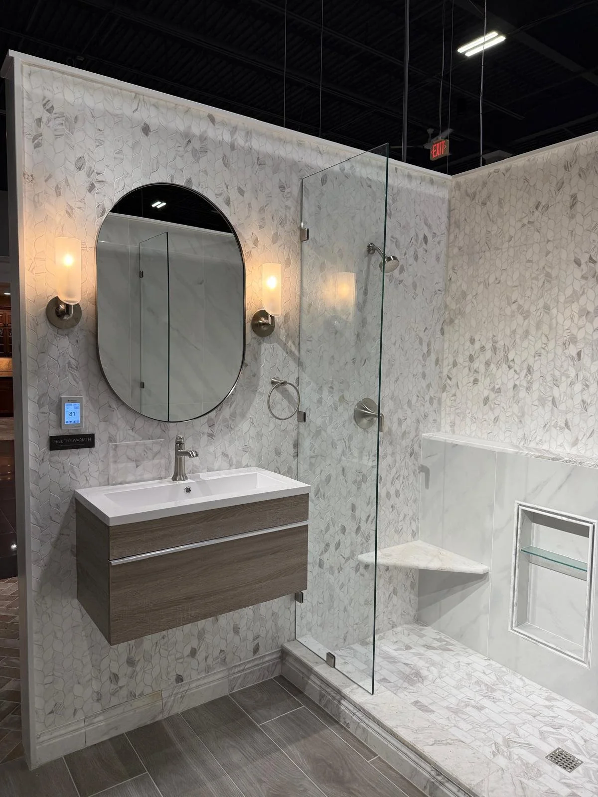 Modern bathroom with a vanity mirror, wall-mounted lights, a floating wooden vanity with a white sink, a digital control panel, and a glass-enclosed shower area with marble and tile surfaces.