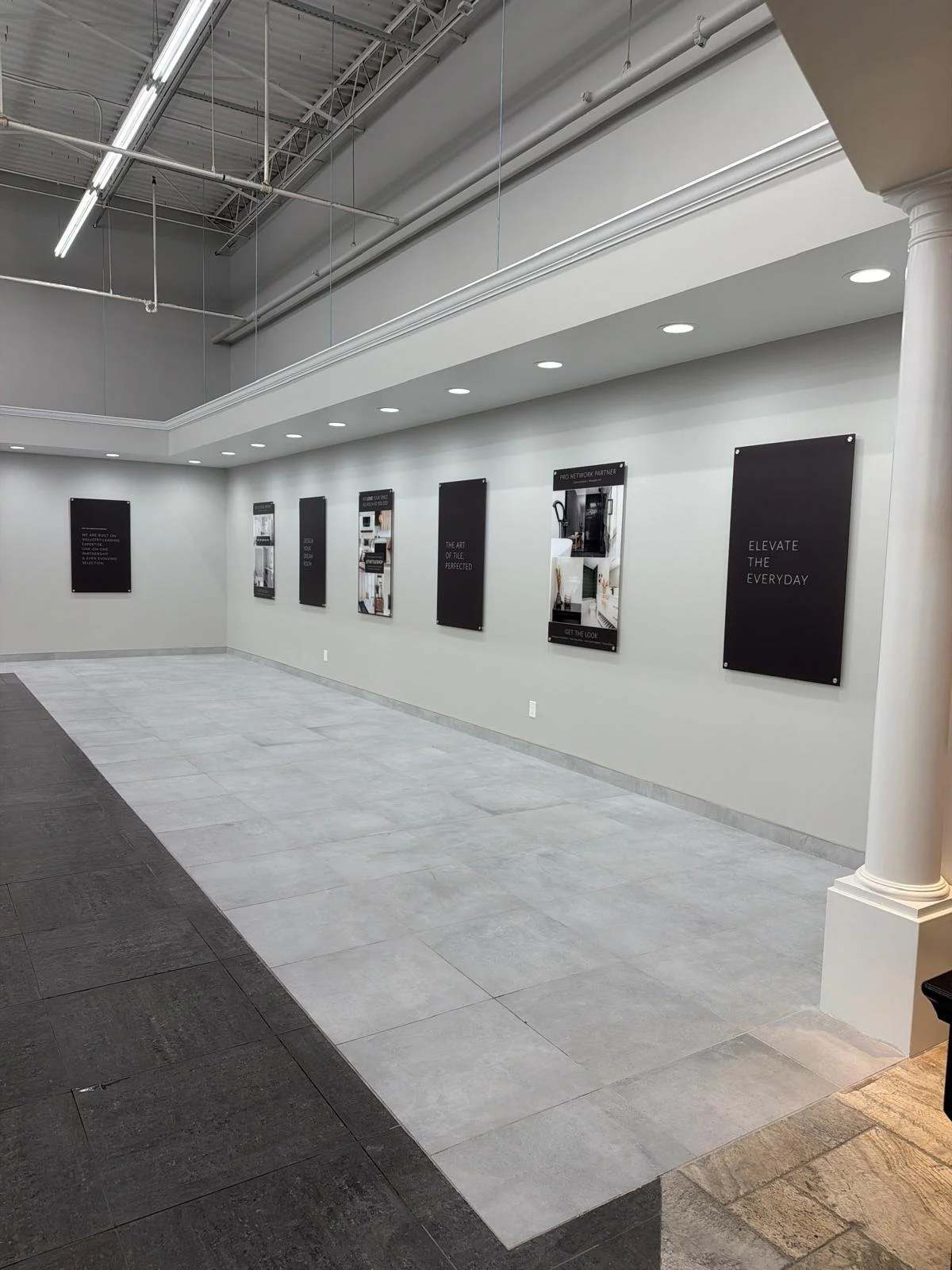 Empty showroom with black and white wall posters, gray tiled floor, white walls, high ceiling with fluorescent lights and exposed piping, white decorative column.