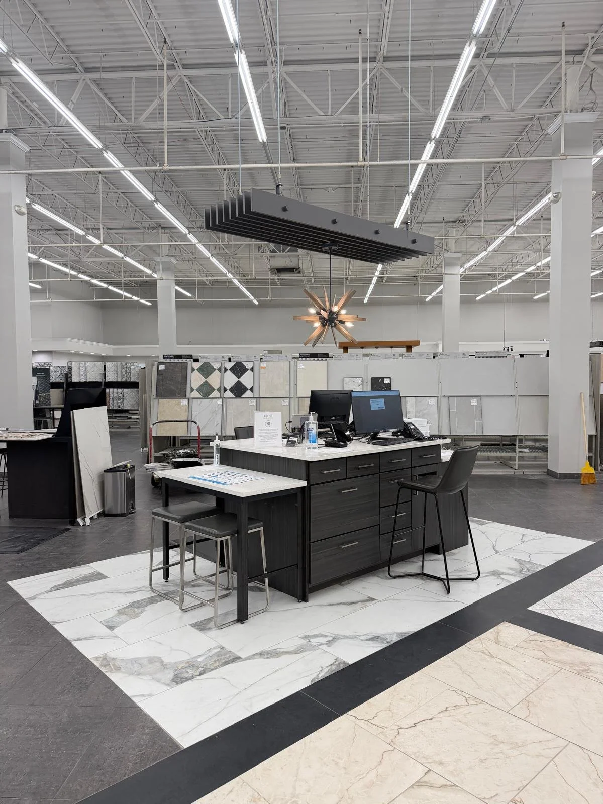 Empty showroom display area with a checkout counter in the foreground, featuring a computer, sanitizer, and chairs. Background shows tile samples on display racks, high ceiling with bright lighting, and a modern light fixture.