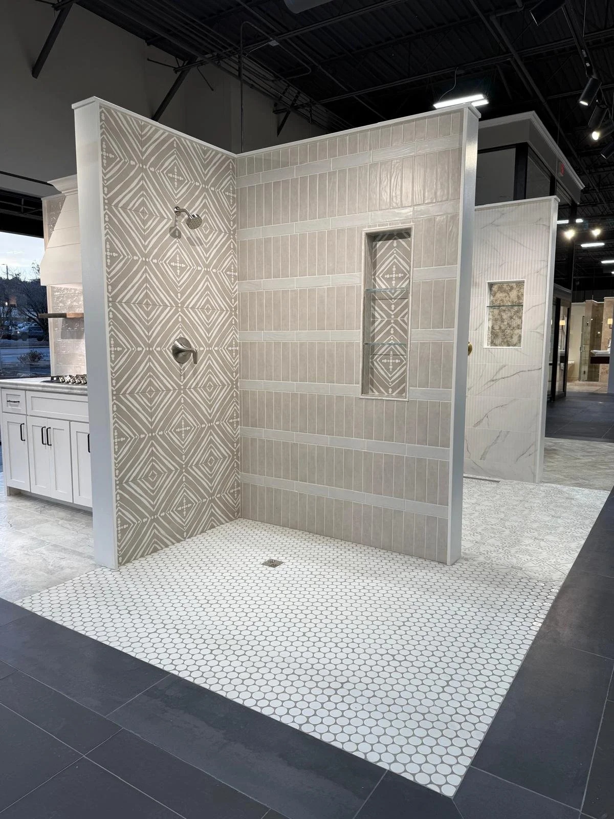 Display of bathroom tiles in a showroom, featuring a shower area with beige geometric patterned wall tiles and a small gray hexagonal tiled floor section, surrounded by other tile displays and dark flooring.