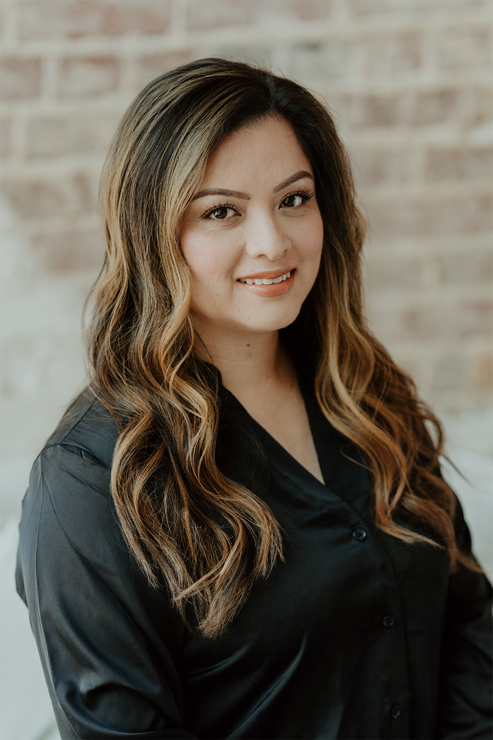 A woman with long, wavy brown hair wearing a black blouse and smiling against a brick wall background.