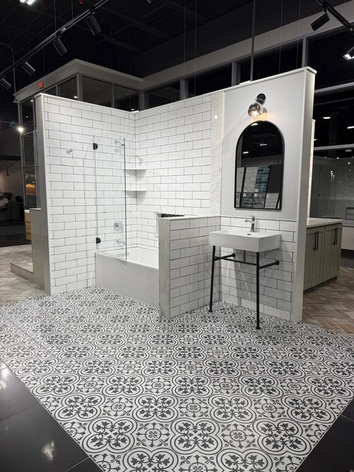 Bathroom display with a white tiled shower with a glass door, a white industrial-style sink with black metal legs, a mirror with a black frame, and intricate black-and-white patterned floor tiles in a showroom setting.