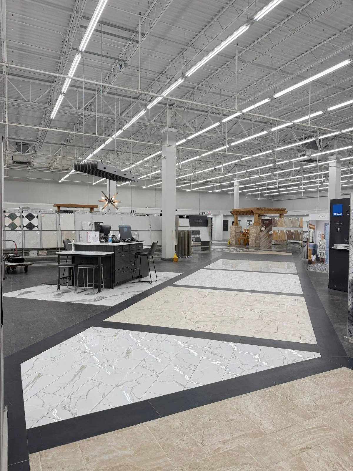 Interior of a tile showroom with display samples on the walls and various tile choices on the floor, a checkout counter with computers, and manufacturing or display booths in the background.