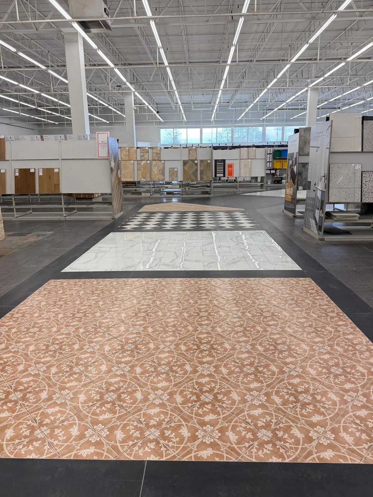 Interior of a flooring store with display samples of different tile and flooring options arranged on metal racks and an aisle with patterned flooring in the foreground, fluorescent lighting, and large windows at the back.