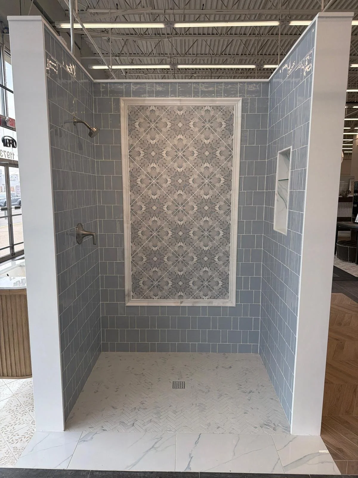A shower display with light blue tiles on the walls, a decorative tile accent on the back wall, a recessed shelf on the right, and a simple school floor drain in the center. The shower also has a showerhead and a control handle on the left wall.