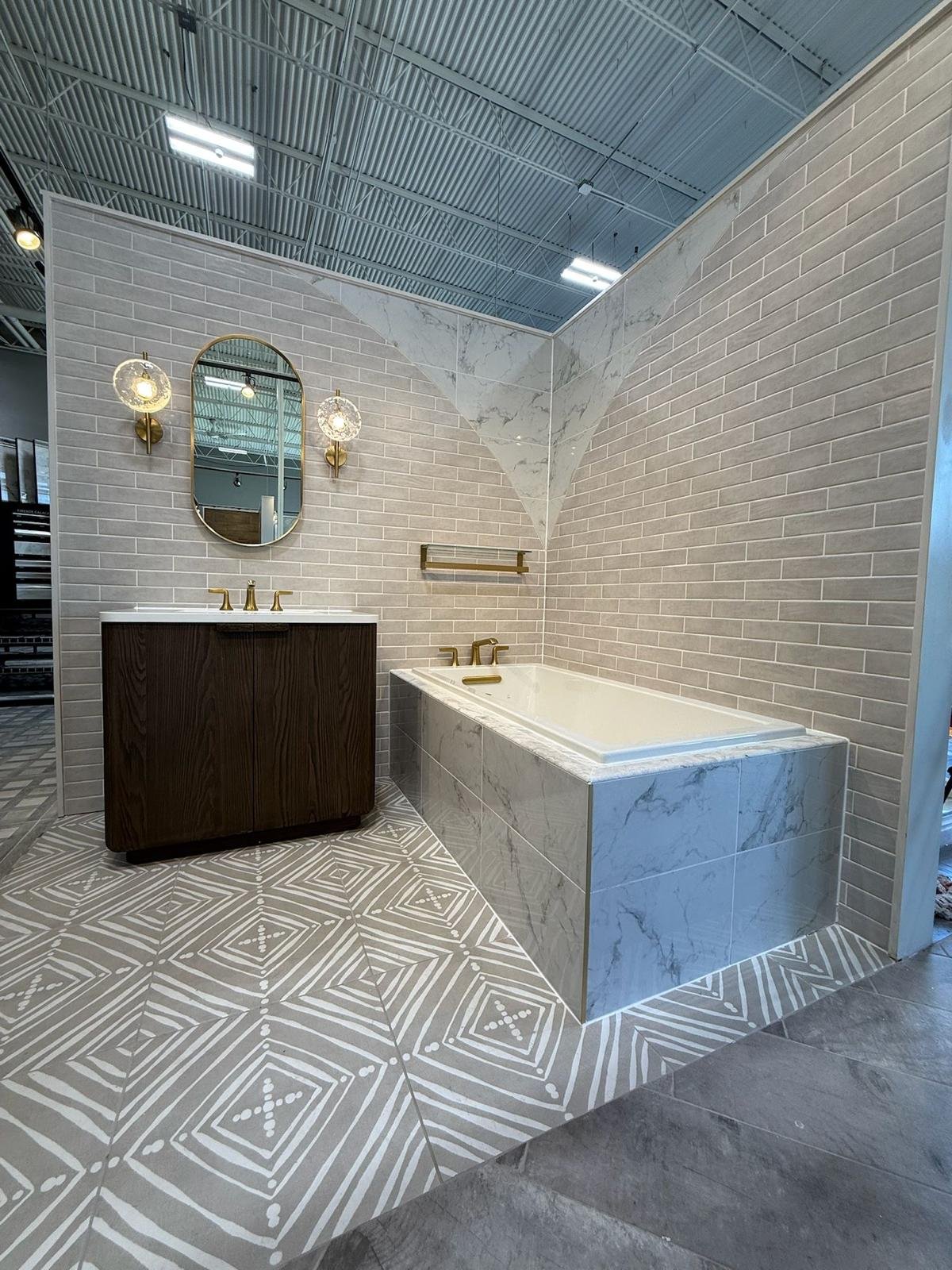 Modern bathroom with a built-in bathtub surrounded by marble tiles, a wooden vanity with a mirror and two wall-mounted light fixtures, beige brick walls, patterned tile flooring, and a wall-mounted towel bar.