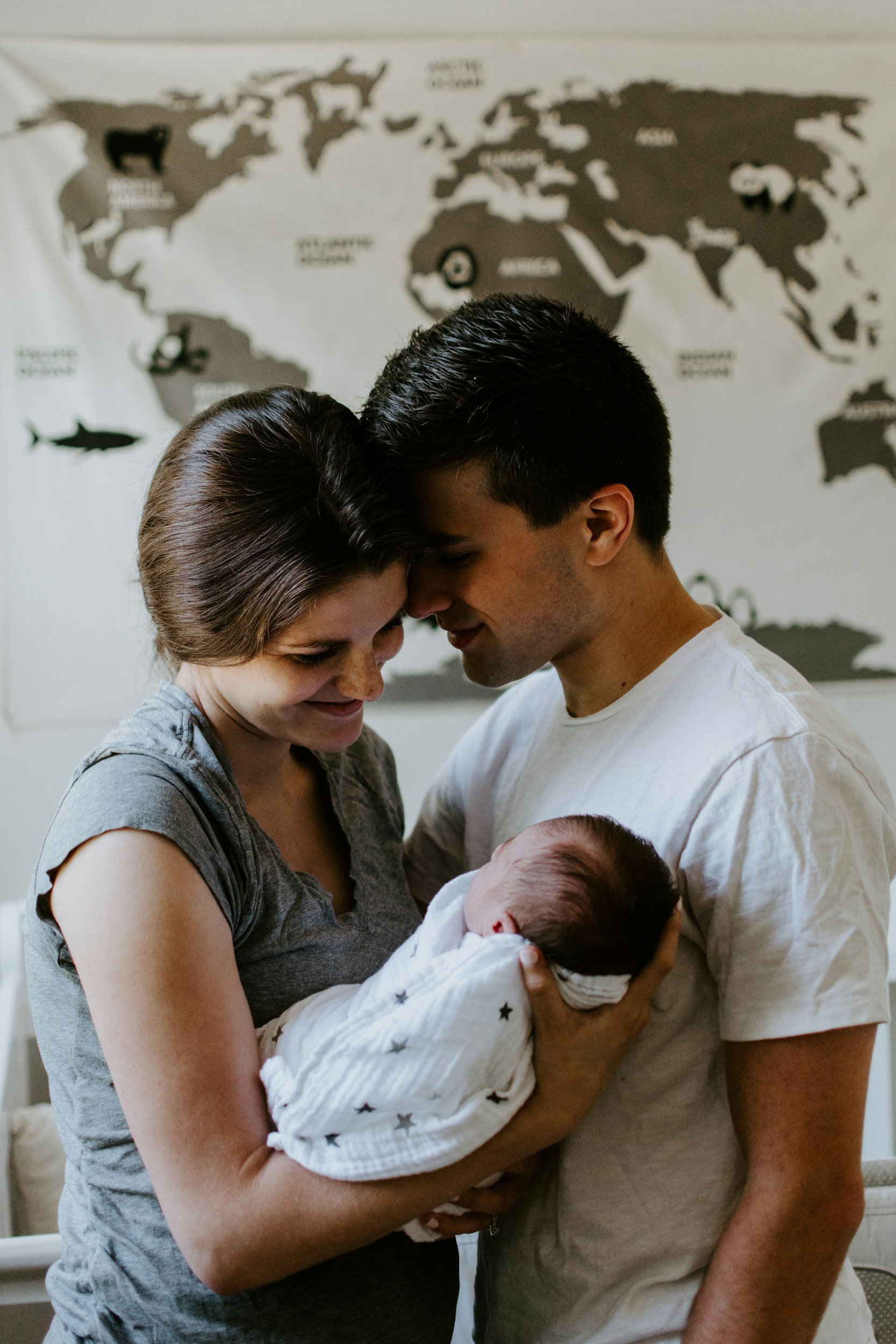 Una coppia che tiene un neonato tra le braccia, in un ambiente domestico, con una mappa del mondo sulla parete sullo sfondo.