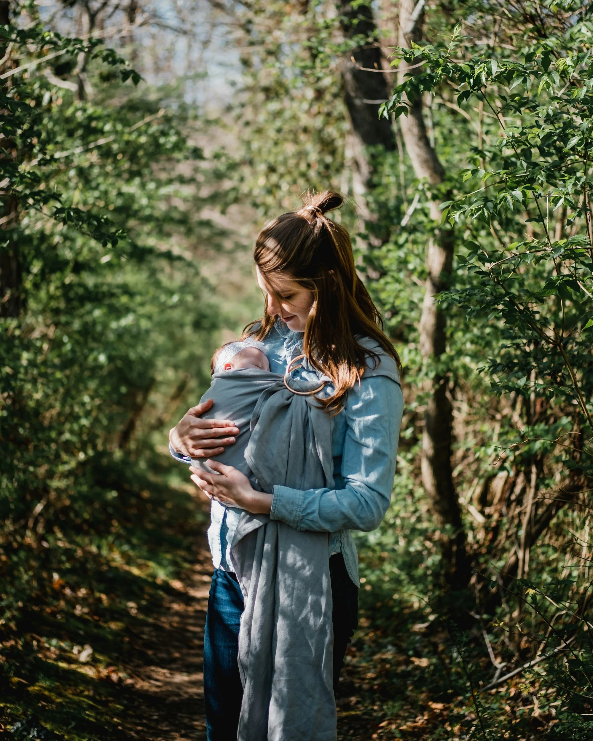 Una donna abbraccia un bambino in un bosco, con alberi e foglie verdi sullo sfondo.
