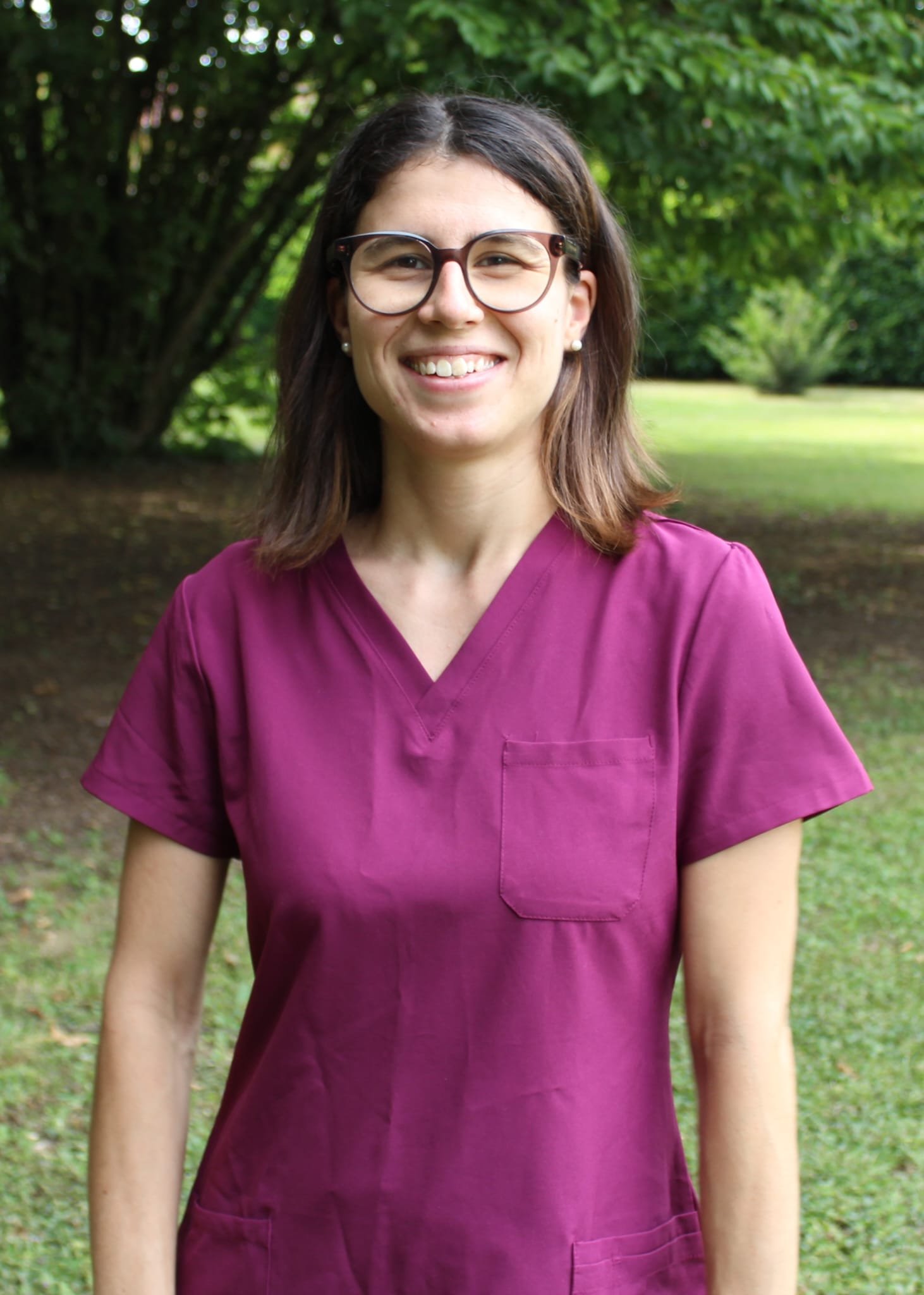 Una donna sorridente con occhiali, indossa una divisa viola ed è all'aperto in un parco con alberi verdi sullo sfondo.