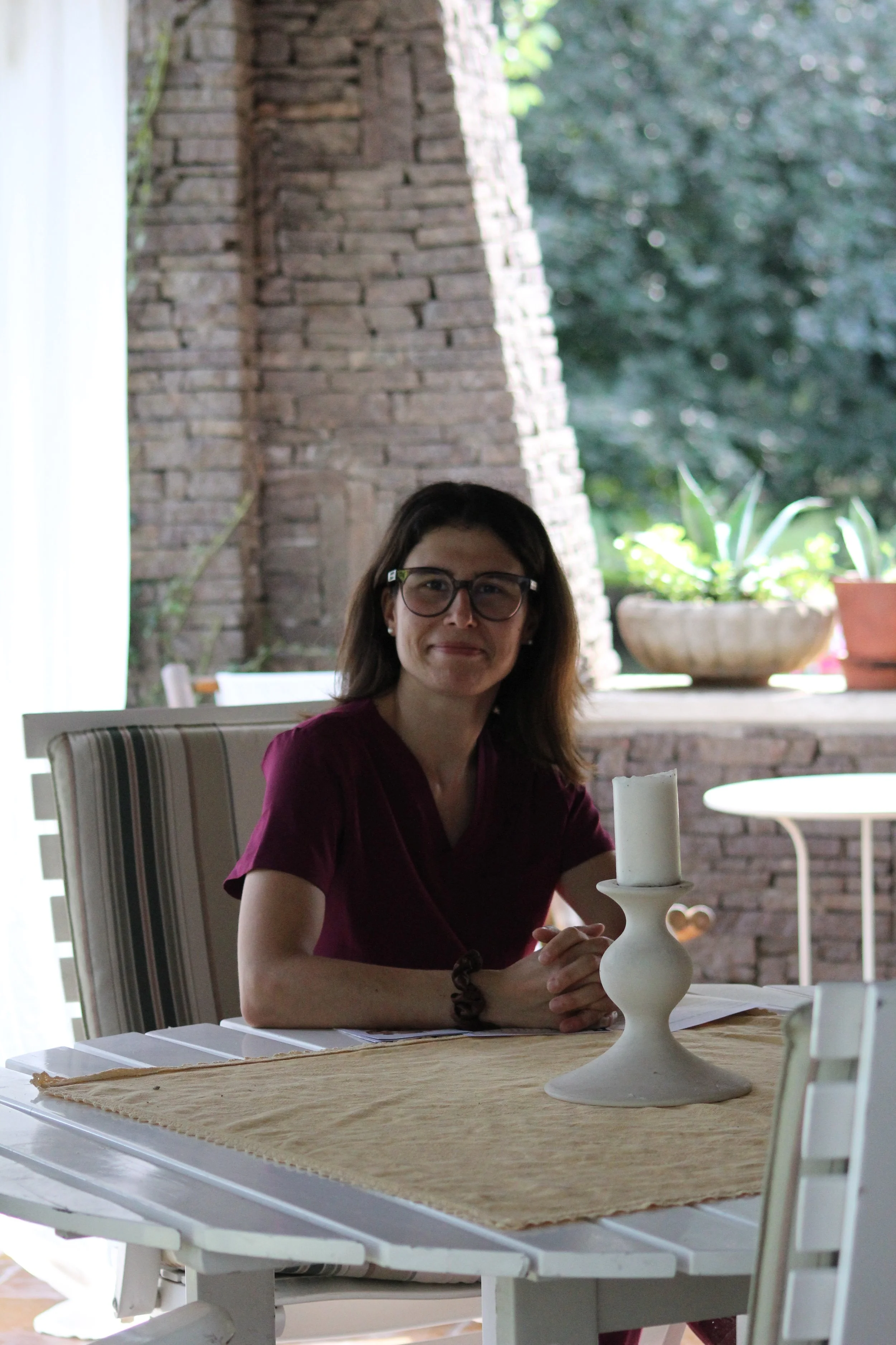 Denise seduta a un tavolo interno con pareti di mattoni e piante sullo sfondo, tiene in mano una candela bianca su un portacandele.