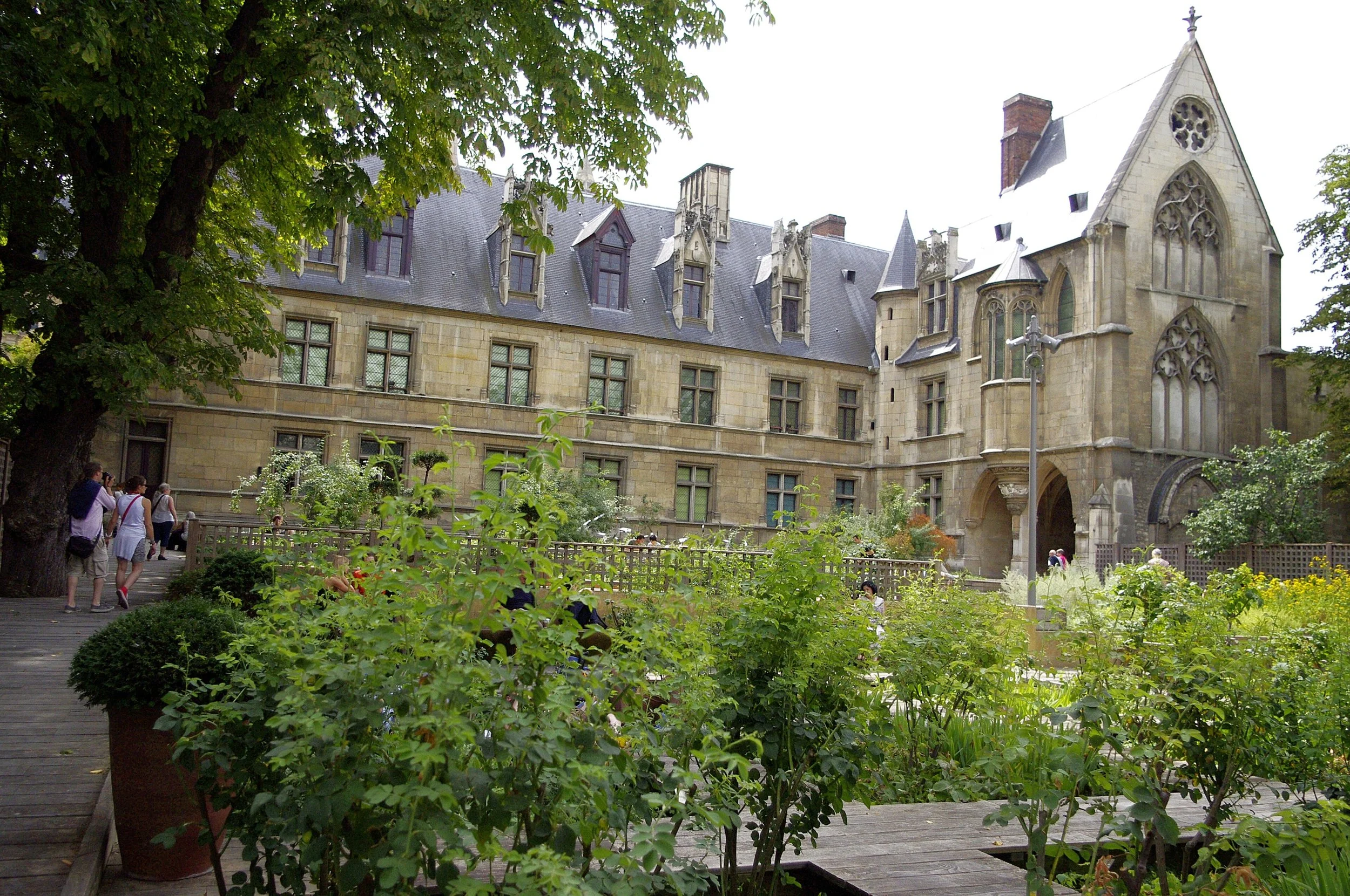 A historic, Gothic-style building with pointed arches, tall windows, and a steeply pitched roof, surrounded by lush greenery and people walking in a garden courtyard. Hôtel de Cluny in Paris, France.