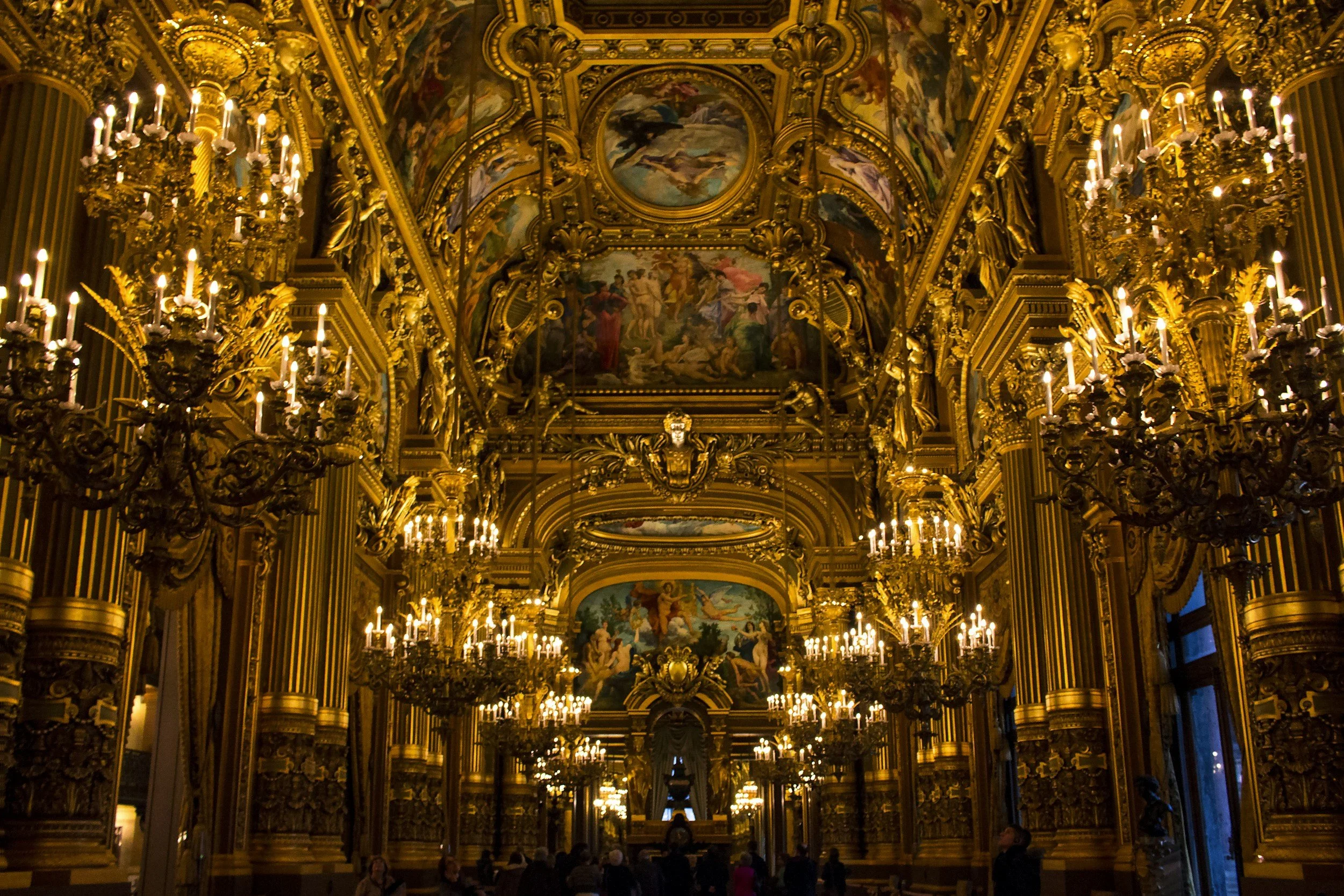 An opulent ornate hall from Opéra Garnier in Paris, decorated with gold accents, large chandeliers, and painted ceilings. People are seated at tables in the dimly lit space.